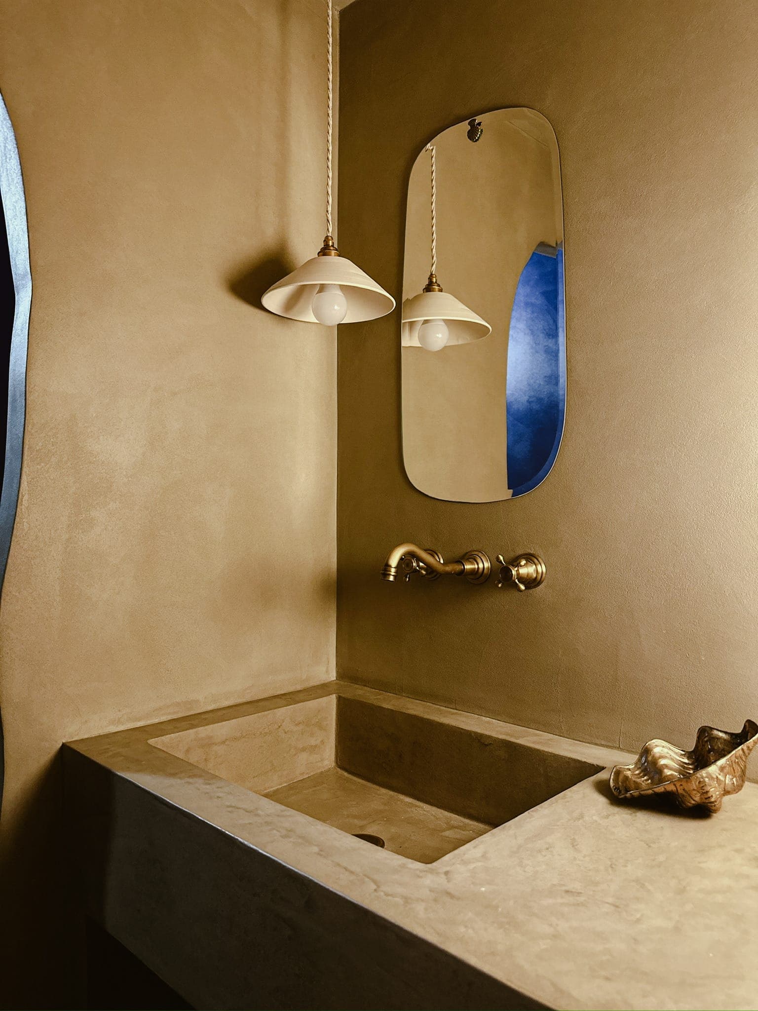 Salle de bain minimaliste avec un lavabo en pierre beige et un robinet en laiton. Au-dessus du lavabo se trouvent un miroir ovale et deux suspensions modernes. Une coquille décorative est posée sur le plan de travail du lavabo. Les murs sont peints dans un ton neutre et chaleureux.