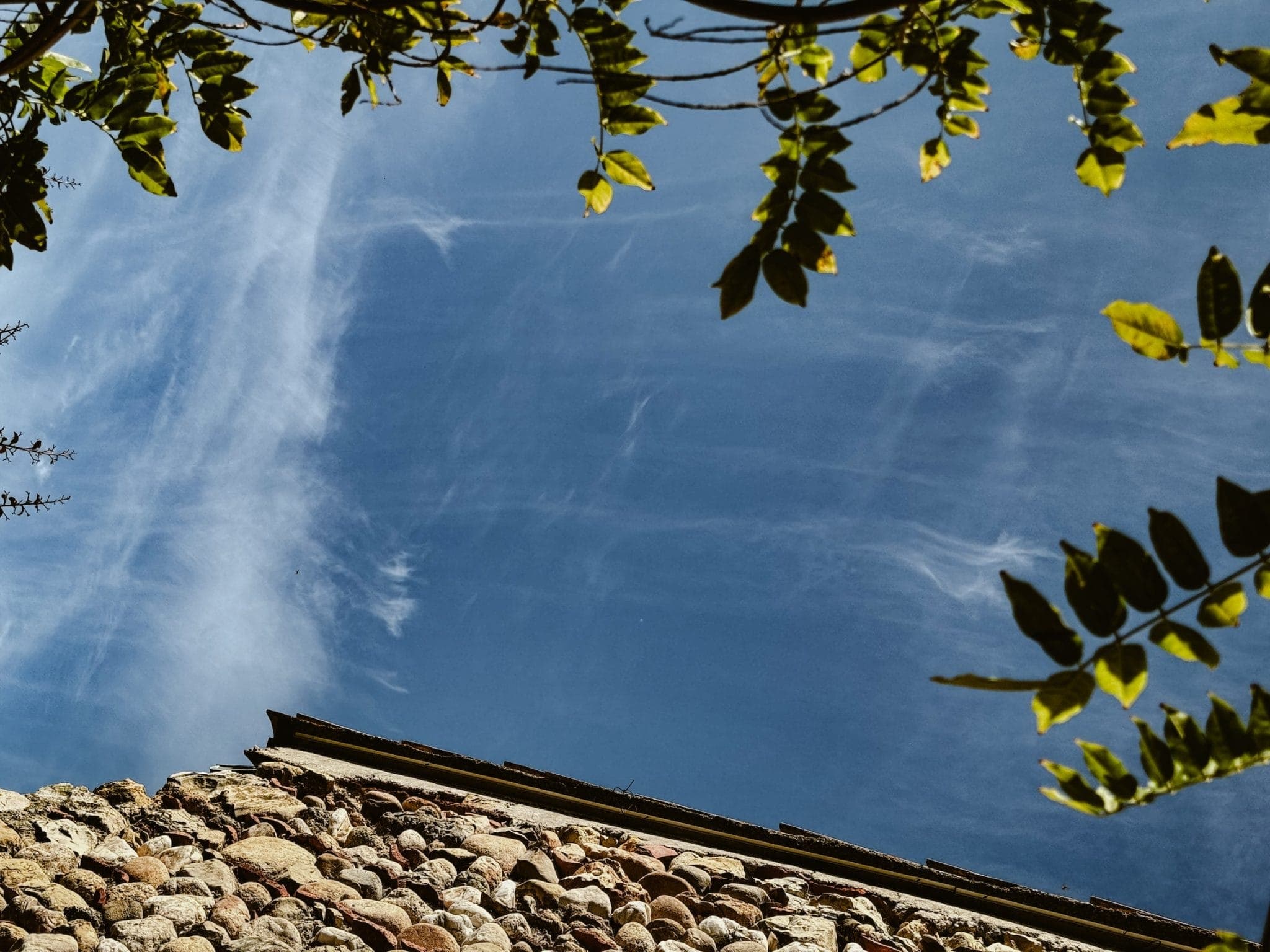 Un mur de pierre rencontre un ciel bleu clair parsemé de nuages fins. Des feuilles vertes encadrent le haut de l'image.