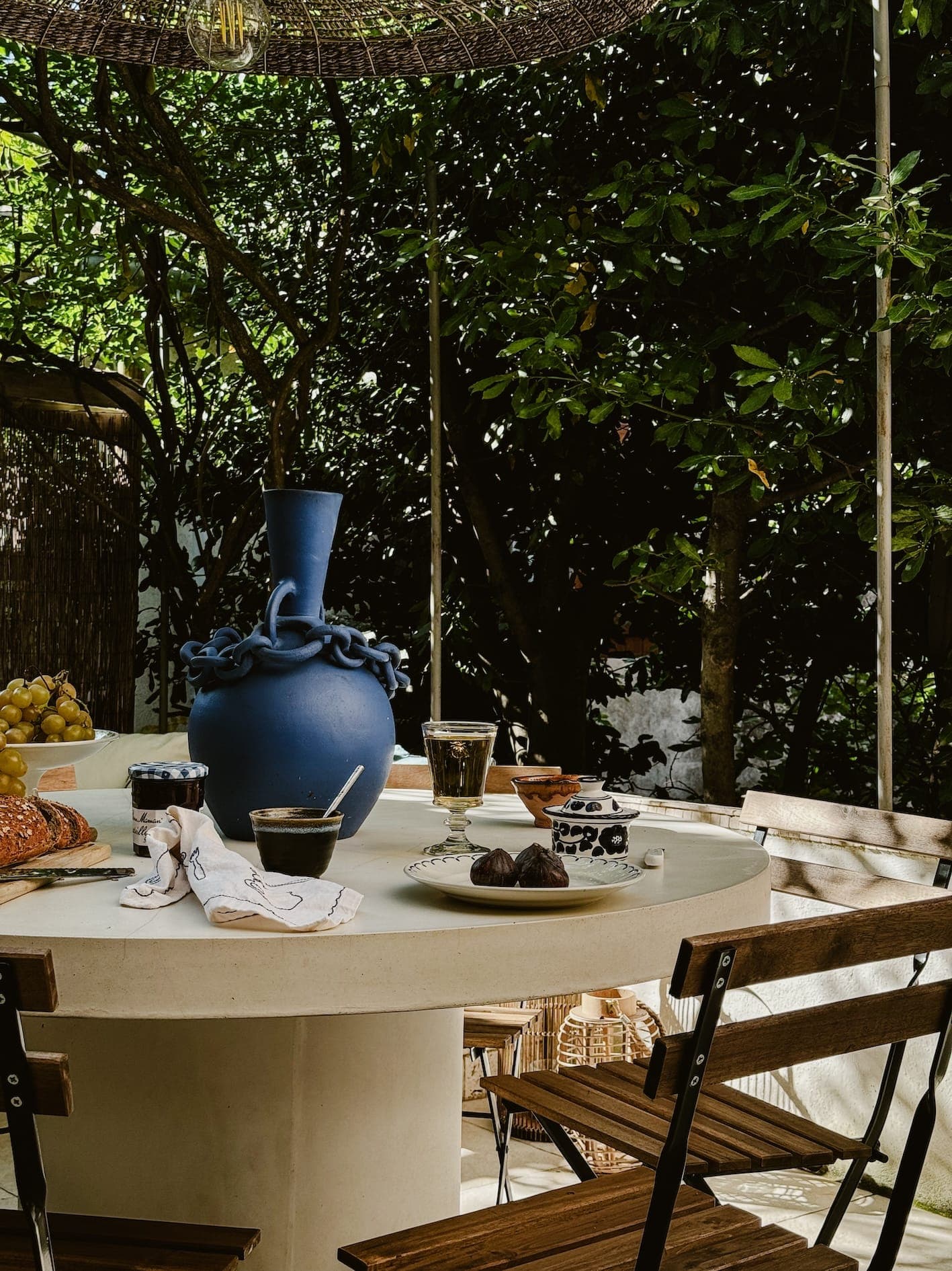 Une table ronde extérieure avec des chaises en bois est dressée pour un repas. Elle comprend un grand vase bleu, des assiettes de nourriture, des verres avec des boissons et un bol de figues. Le cadre est ombragé par des arbres et entouré d'une végétation luxuriante.