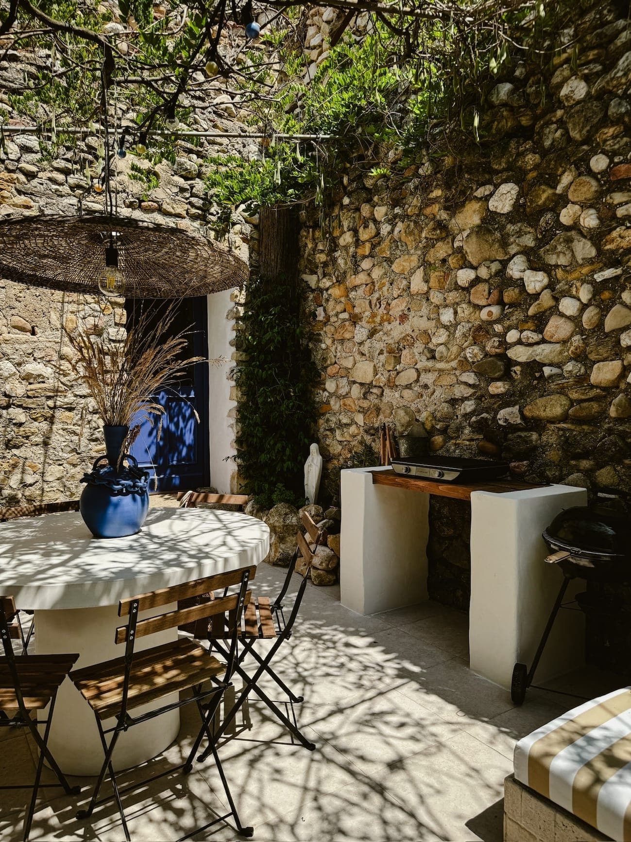 Une terrasse ensoleillée aux murs en pierre abrite une table ronde avec des chaises en bois, surmontée d'un vase bleu rempli de plantes séchées. Une lampe en osier projetant de l'ombre est suspendue au-dessus. Une petite cuisine extérieure avec un barbecue et un comptoir est installée contre le mur, entourée de verdure.