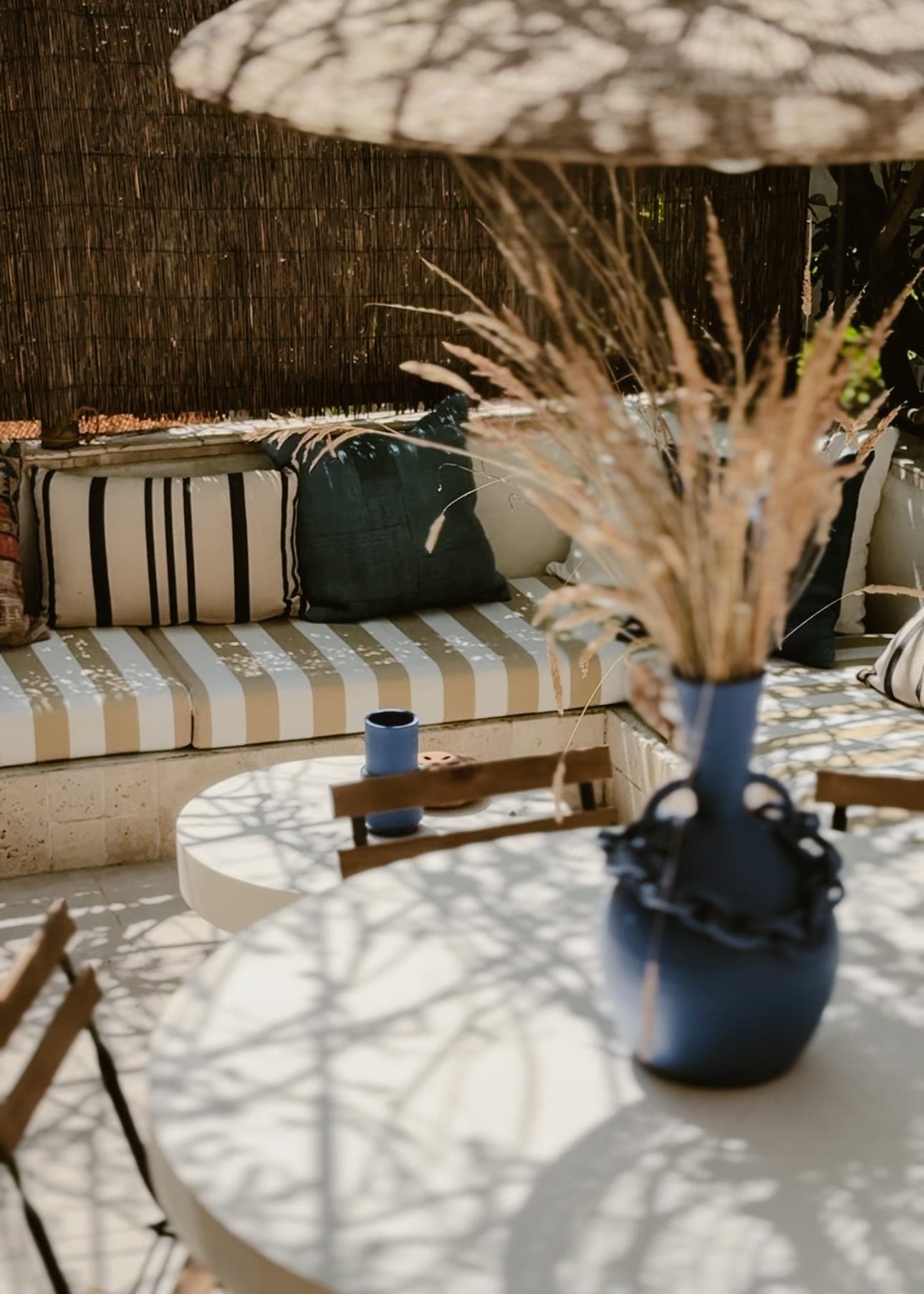 Scène de terrasse extérieure avec un banc rembourré à rayures orné de coussins, une table ronde et un vase bleu contenant des herbes séchées. Un parasol en paille projette des ombres, créant une atmosphère chaleureuse et ombragée.