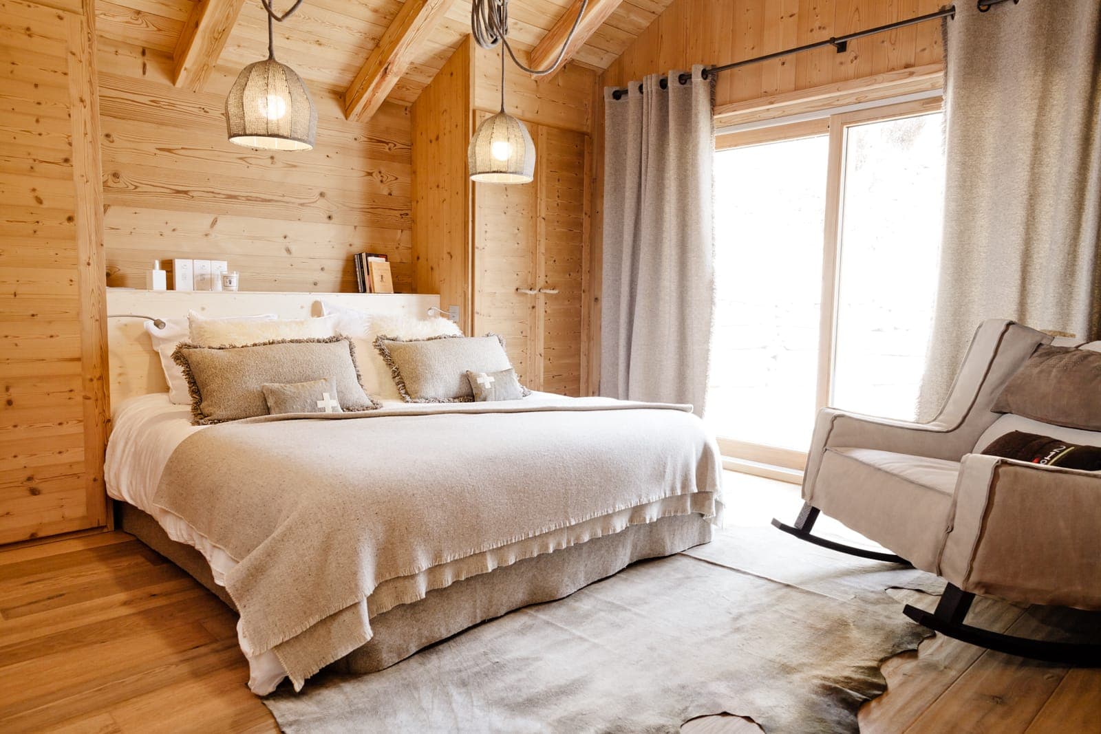 Cozy bedroom with wooden walls and ceiling, featuring a large bed with beige bedding and pillows. A soft rug is on the wooden floor. A rocking chair is beside the window, which is dressed with long gray curtains. Two pendant lights hang above the bed.