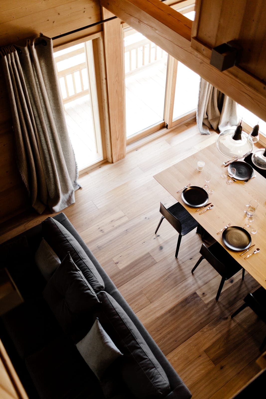 A cozy wooden interior with a dining table set with black plates and cutlery, a dark gray sofa, and large windows with light curtains. Sunlight streams in, highlighting the natural wood flooring and walls, creating a warm and inviting atmosphere.