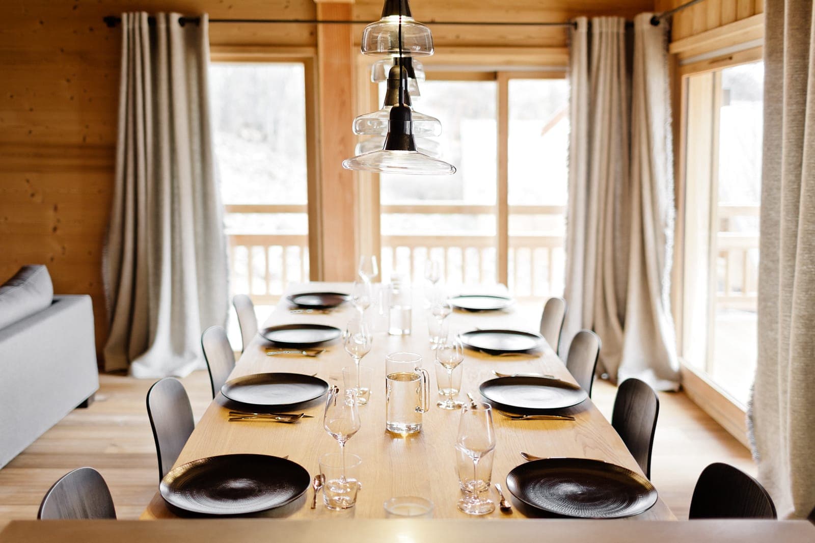 A cozy dining room features a long wooden table set with black plates, wine glasses, and clear pitchers. Modern pendant lights hang above, and large windows with curtains allow natural light to fill the space, highlighting the wooden interior.