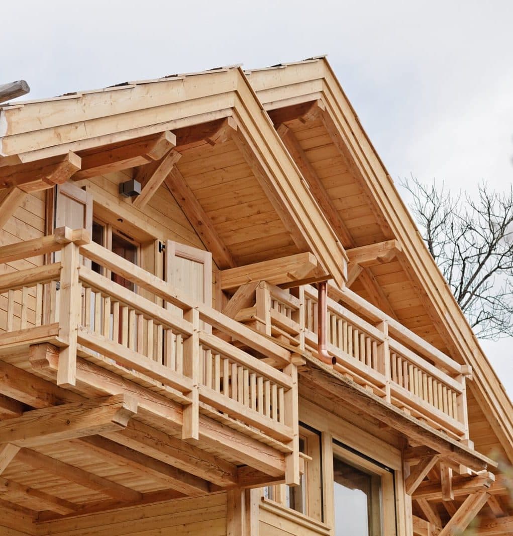 Un chalet en bois avec balcon est représenté sur un fond d'arbres sans feuilles. Le ciel est couvert et les détails de la construction en bois sont visibles. Quelques branches nues sont au premier plan.
