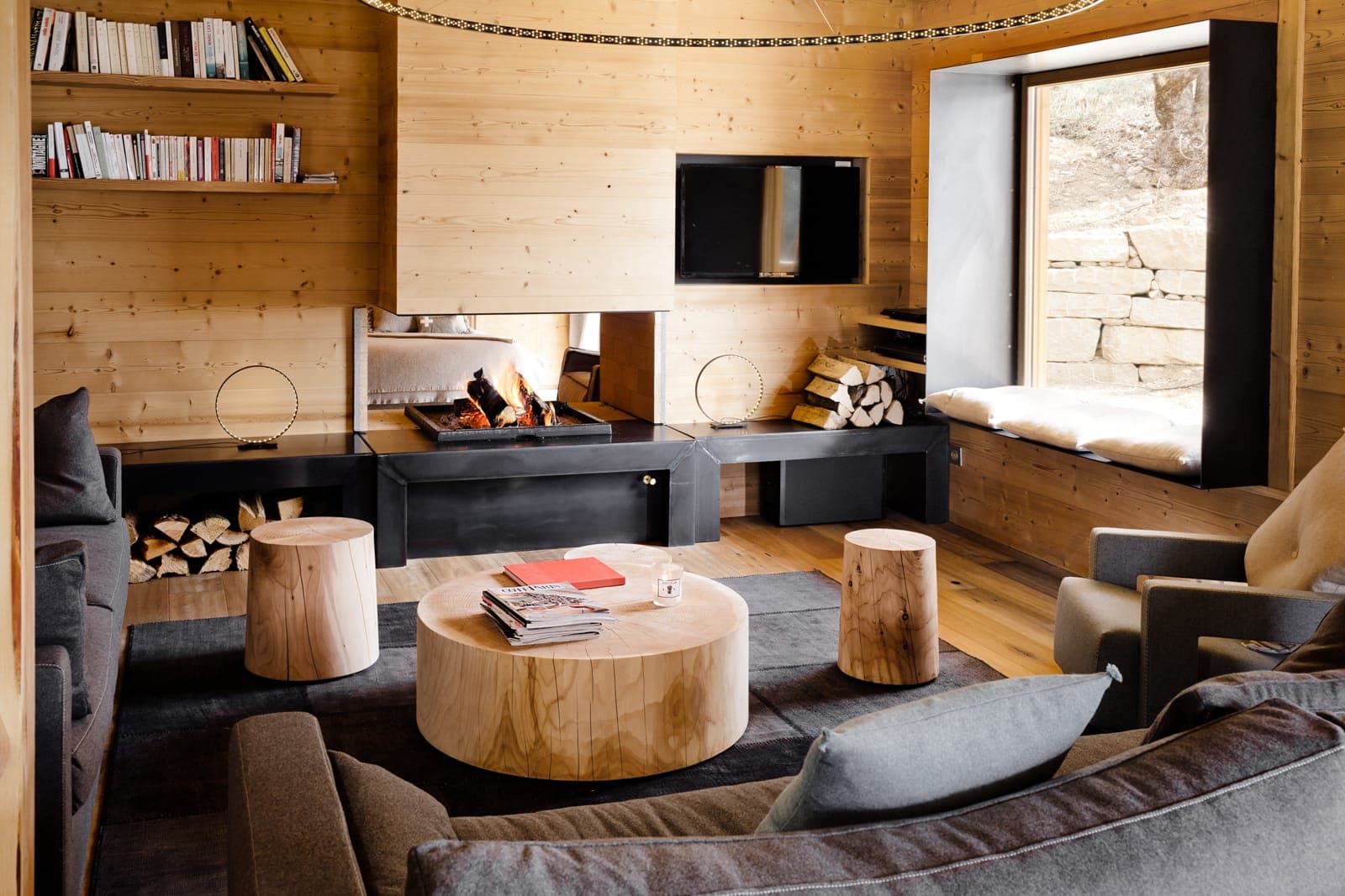 A cozy living room with wooden walls and floor, featuring a fireplace with stacked logs, shelves with books, and a large window. There are comfortable gray sofas and a round wooden coffee table in the center.