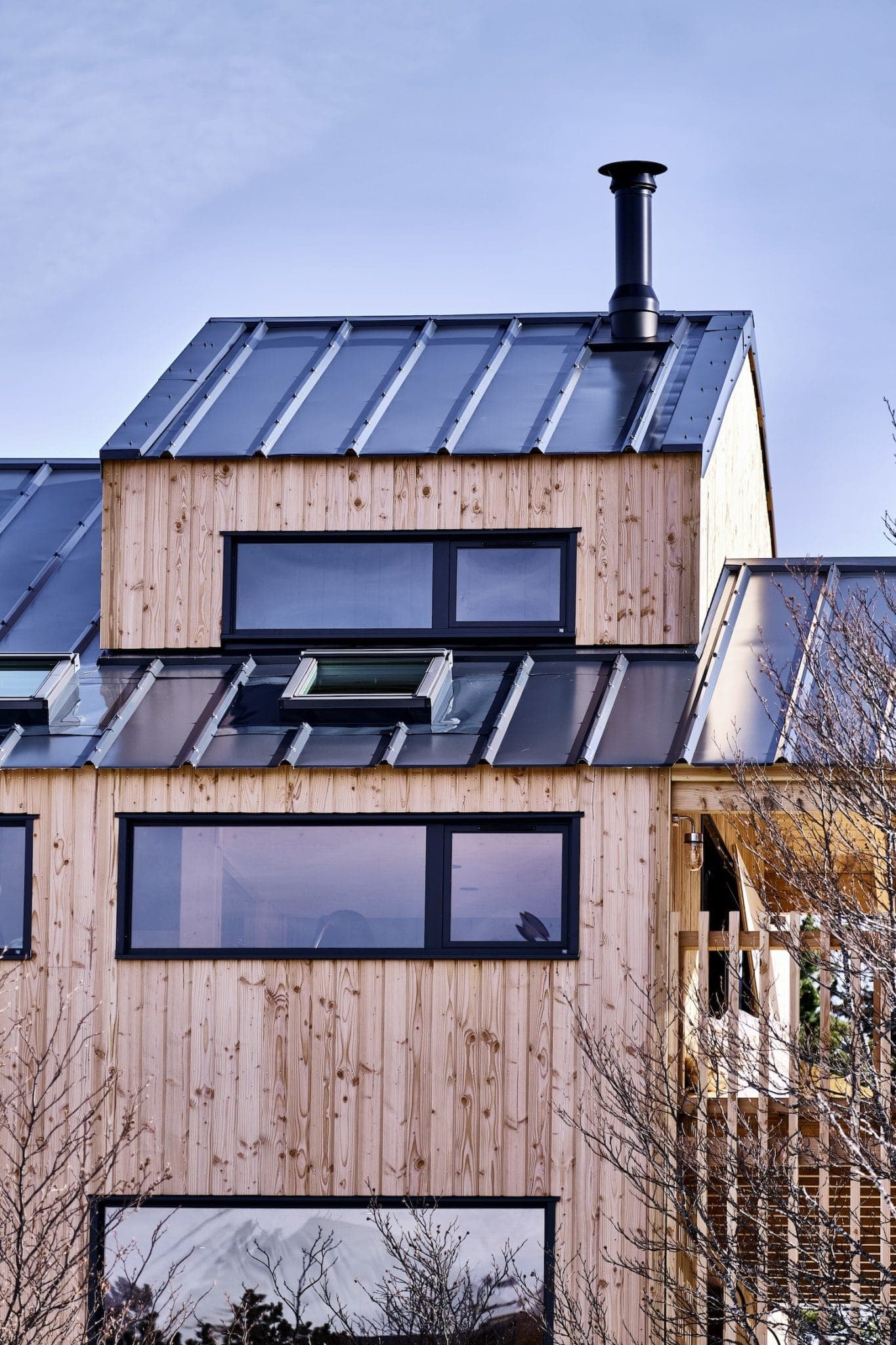 Maison moderne en bois avec de grandes baies vitrées et un toit en métal avec cheminée. La conception intègre des lignes de toit inclinées et une façade en bois naturel, sur fond de ciel bleu clair avec des branches clairsemées au premier plan.