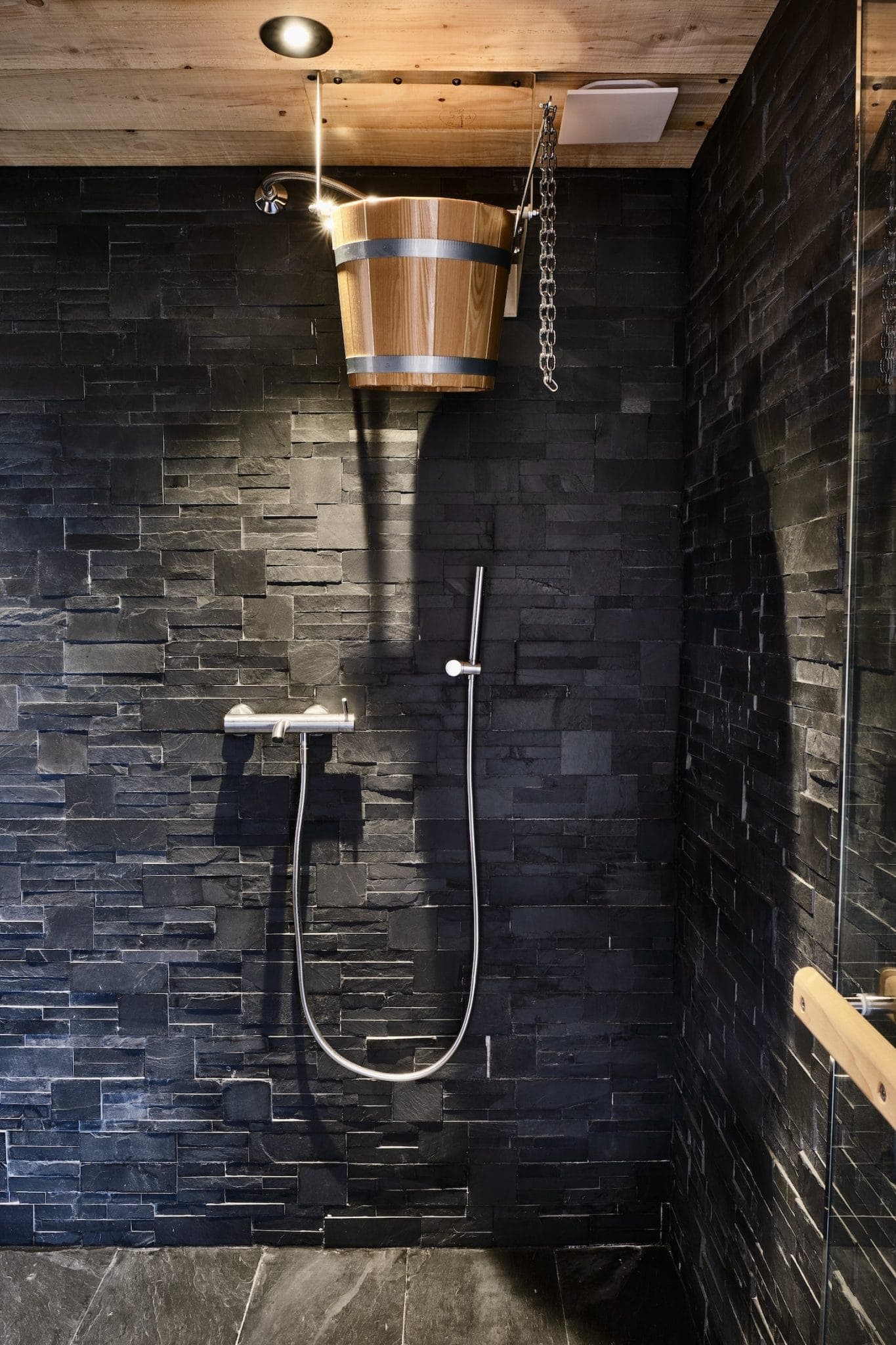 Une douche moderne avec des murs en carreaux de pierre foncée, avec une pomme de douche à effet pluie et un seau d'eau en bois suspendu au-dessus, relié par une chaîne. La zone est chaleureusement éclairée par le haut.