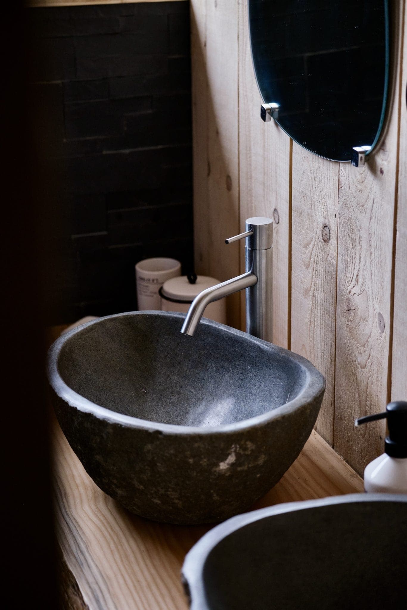 Une salle de bains rustique est dotée d'un lavabo en pierre avec un robinet en métal élégant, posé sur un plan de travail en bois. Un miroir rond avec un cadre sombre est suspendu au-dessus et divers articles de toilette sont placés à proximité. Le mur est fait de panneaux de bois de couleur claire.