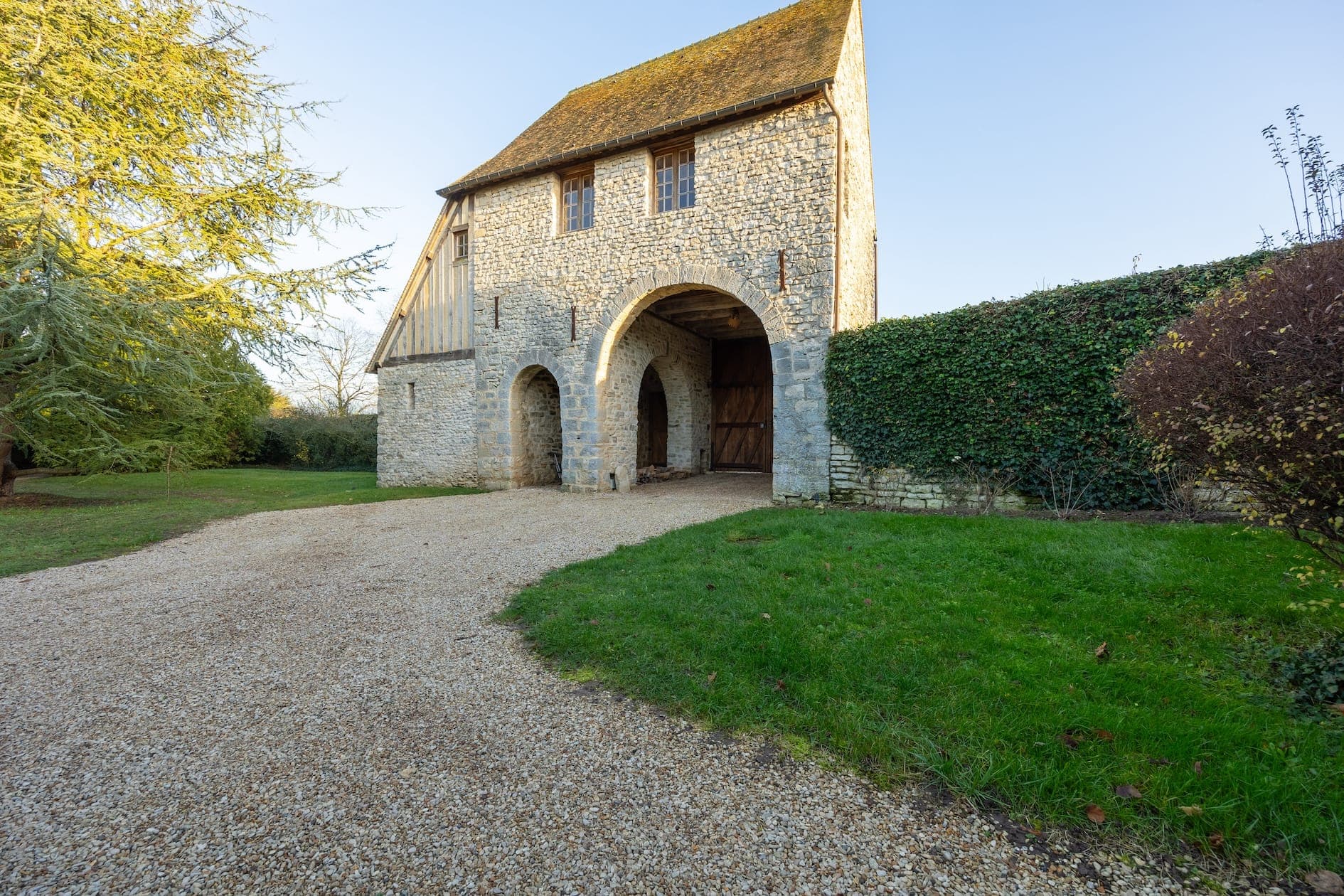 Un bâtiment rustique en pierre avec une entrée voûtée et un toit en pente, situé sur un chemin de gravier. Il est entouré de verdure, notamment d'une pelouse, de haies et d'arbres, sous un ciel bleu clair.