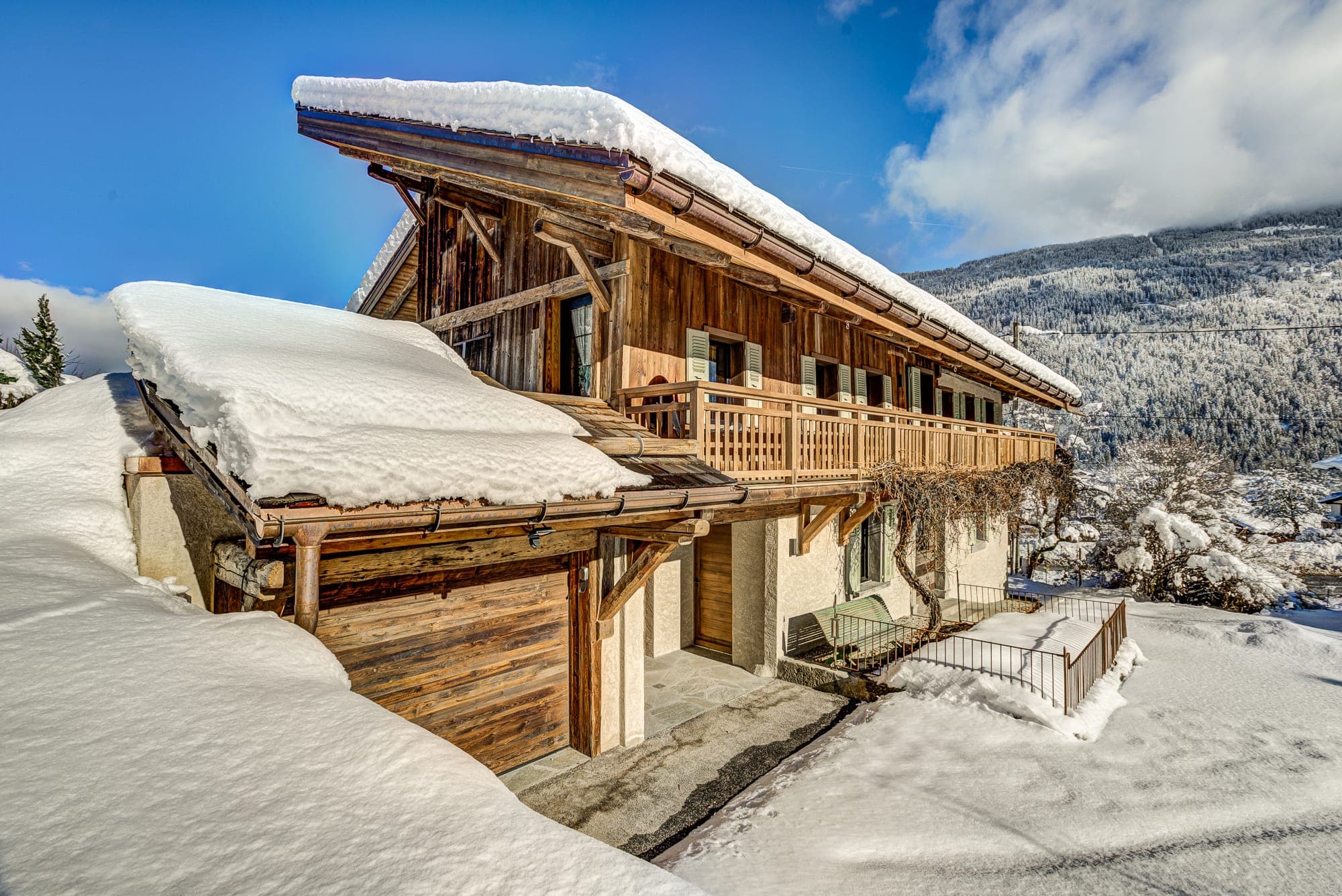 A wooden chalet covered in snow sits amidst a snowy landscape. The sky is clear and blue, with a few clouds, and a mountain is visible in the background. The scene is serene and picturesque.