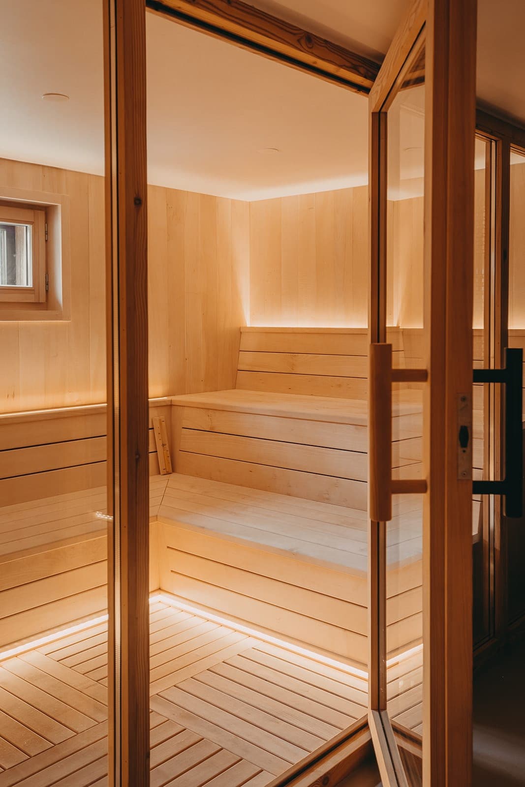 Un sauna en bois confortable avec un éclairage d'ambiance chaleureux. La pièce est dotée de bancs en bois à plusieurs niveaux et d'une porte vitrée, créant une atmosphère sereine et accueillante. Une petite fenêtre près du plafond laisse entrer une douce lumière naturelle.