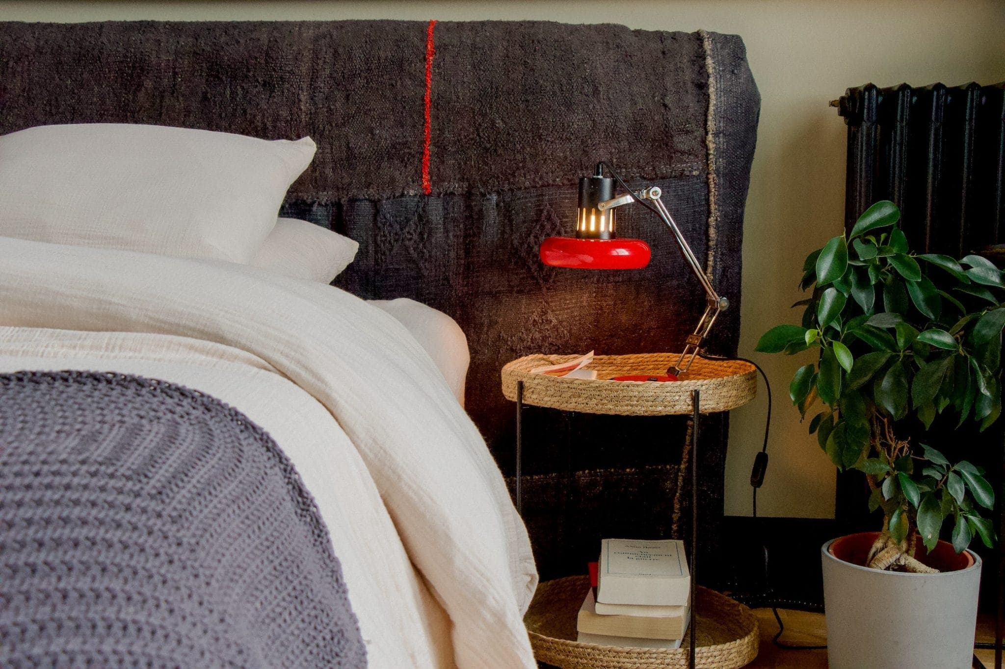 Une chambre douillette avec un lit bien fait, des oreillers blancs et une couverture grise. Une petite table d'appoint ronde abrite des livres et une lampe de bureau rouge. Une plante en pot est à proximité, ajoutant une touche de verdure.