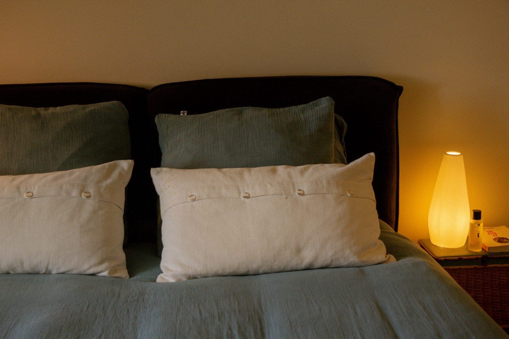 Un lit bien fait avec deux gros oreillers et deux petits coussins blancs. Le lit est doté d'une couette bleu-vert. Une lampe douce et chaleureuse ainsi que quelques petits objets reposent sur la table de nuit à droite.