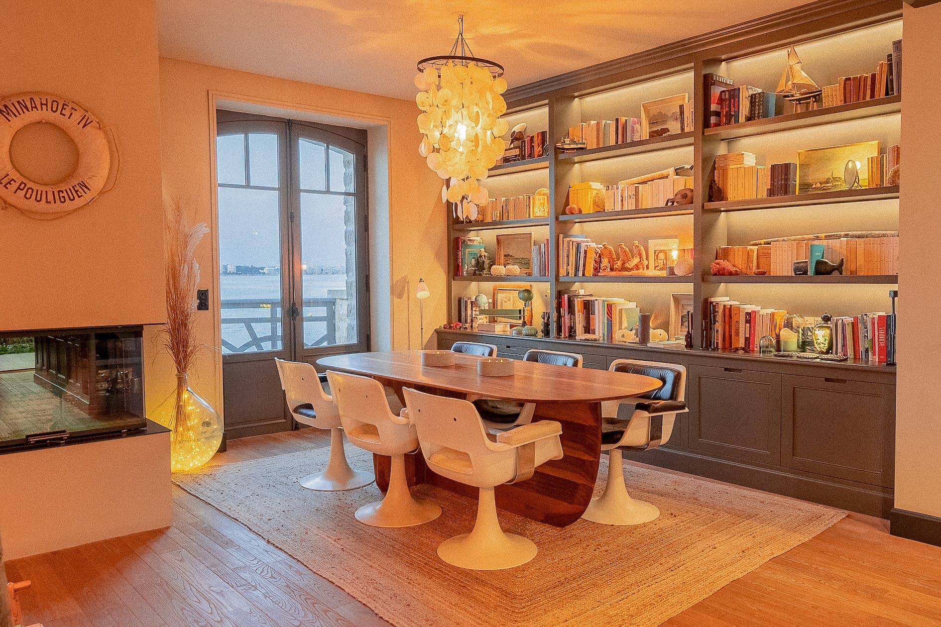 Une salle à manger confortable avec une table en bois et huit chaises, entourée de hautes étagères remplies de livres et de décorations. Un lustre circulaire est suspendu au-dessus. Une grande fenêtre offre une vue sur l'extérieur et une lampe d'angle allumée ajoute de la chaleur à l'espace.