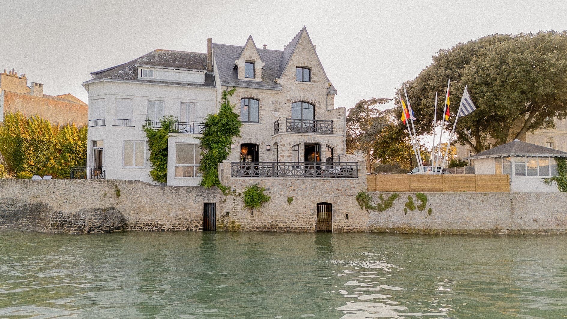 A stone house with large windows and balconies overlooks a calm body of water. Flags and trees are nearby, and the setting sun casts a warm glow on the scene.