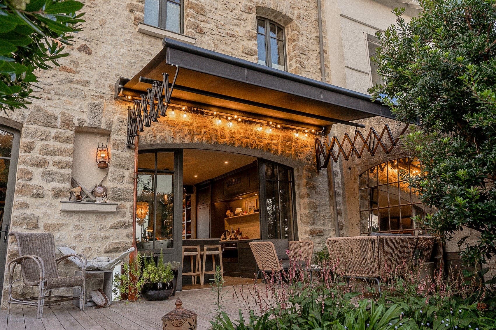 Une terrasse en pierre confortable avec un auvent rétractable est éclairée par des guirlandes lumineuses. Elle dispose de chaises en osier et de petites tables, entourées d'une végétation luxuriante. Les portes coulissantes ouvertes offrent un aperçu d'un intérieur accueillant avec des tabourets de bar et un éclairage chaleureux.