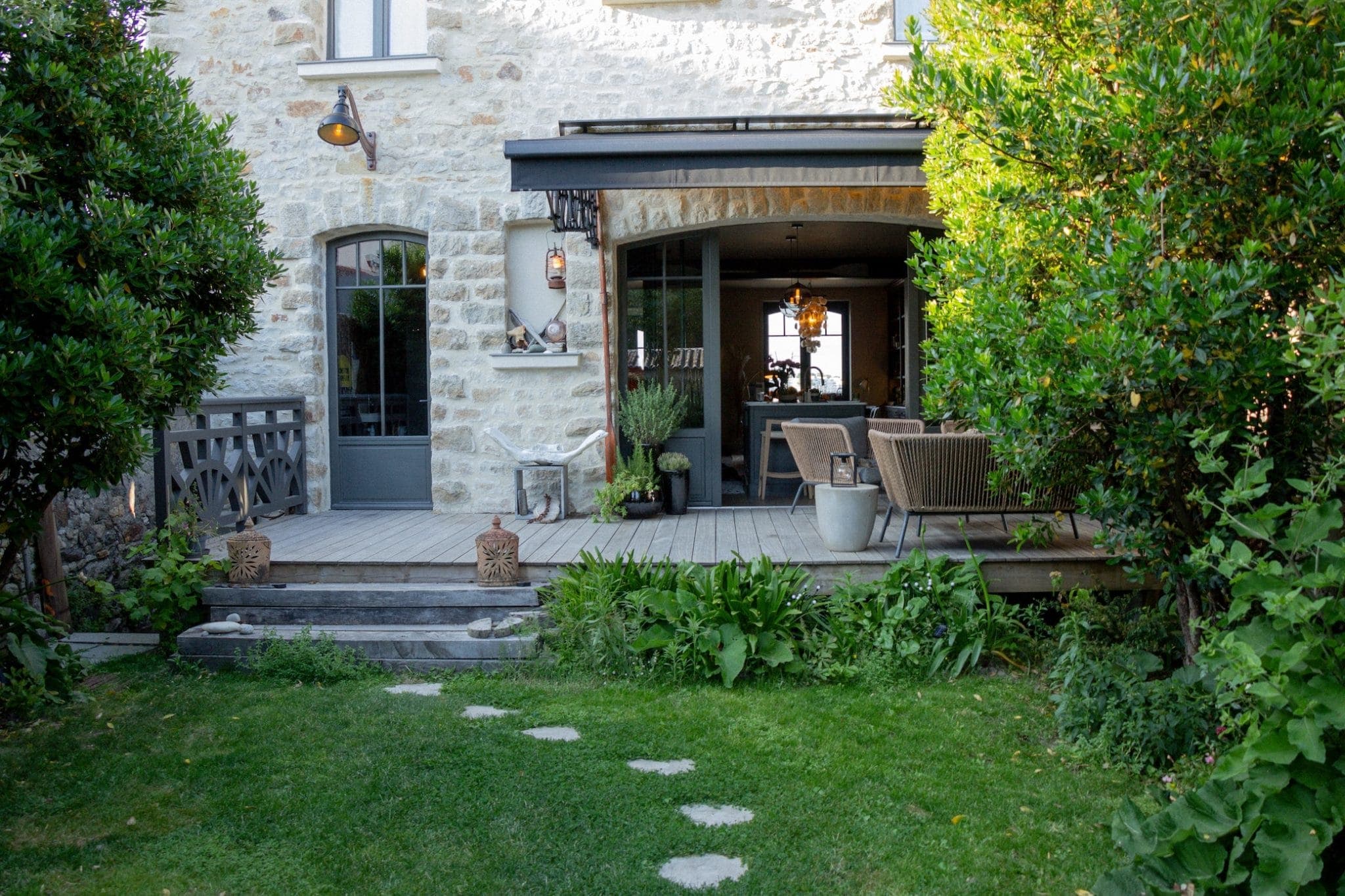 A cozy patio area with wooden chairs and a table is attached to a rustic stone house. Green shrubs and trees surround the patio, and stepping stones lead across the grass. Warm lighting and potted plants add charm to the outdoor space.