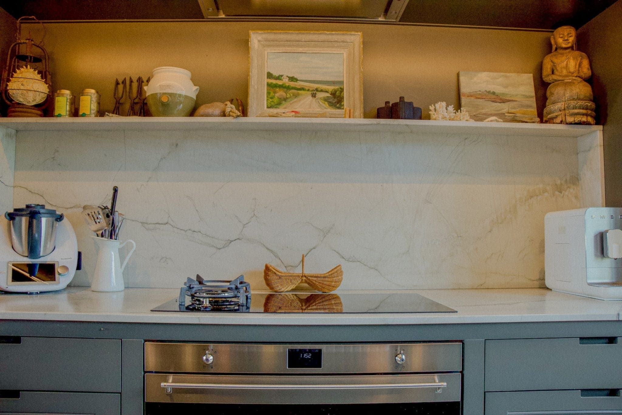 Cuisine moderne avec dosseret en marbre, cuisinière à poser et four. Une étagère au-dessus accueille des objets décoratifs comme un tableau, des bols et une statue. Les appareils de cuisine reposent sur le comptoir de chaque côté.