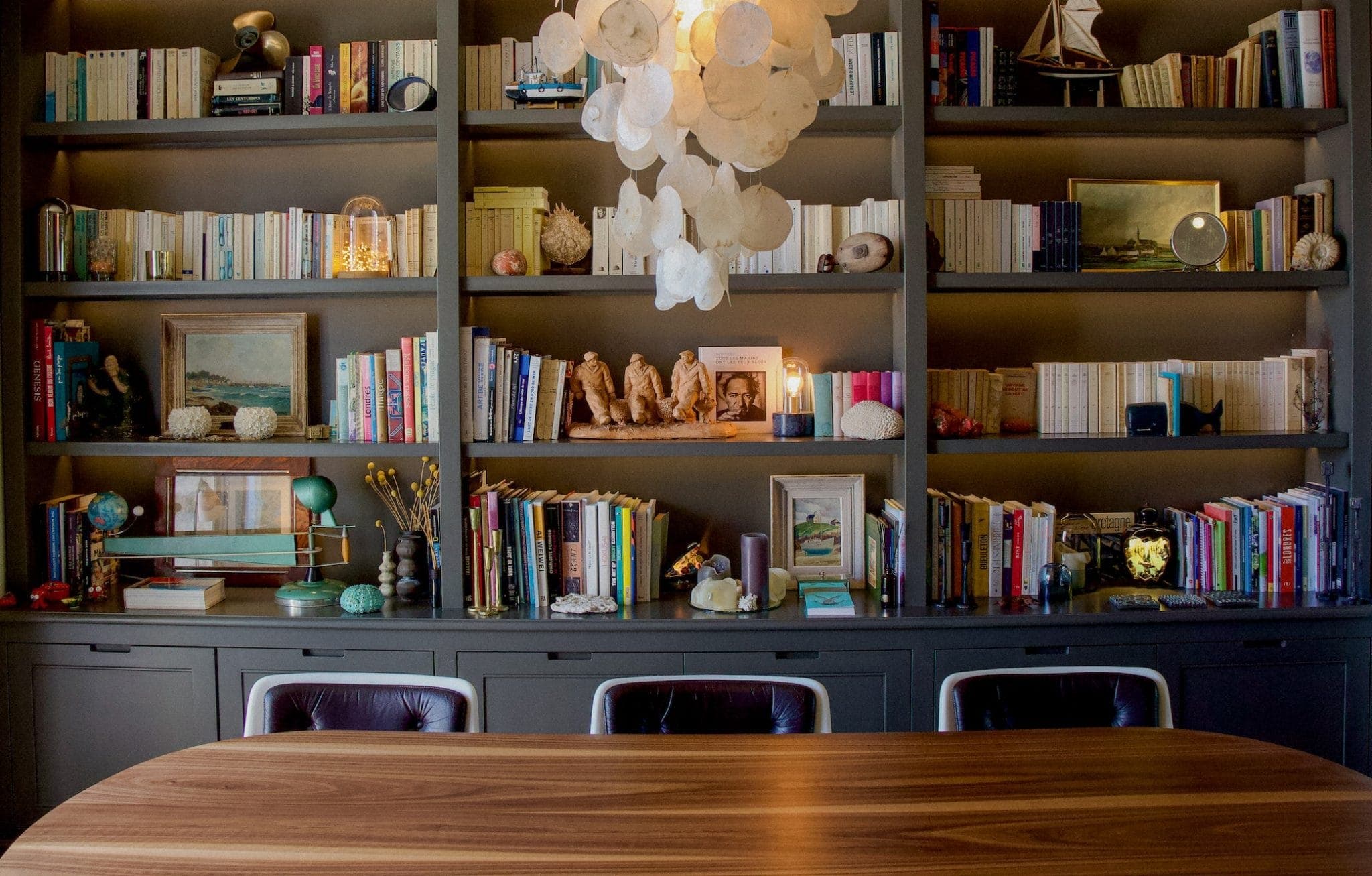 Une pièce chaleureusement éclairée dispose d'une table en bois entourée de chaises devant une grande bibliothèque bien garnie. Les étagères présentent un assortiment de livres, de décorations et de petites œuvres d'art. Un lustre unique avec des éléments circulaires est suspendu au plafond.