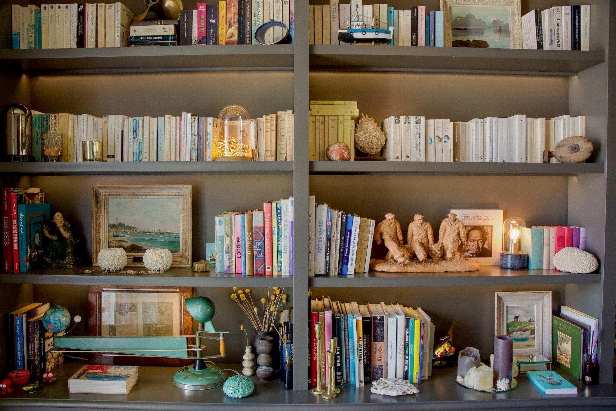Une bibliothèque bien organisée, remplie d'une variété de livres, d'objets décoratifs et d'œuvres d'art. Les articles comprennent un globe, de petites sculptures, des peintures encadrées et une lampe, ajoutant une touche éclectique et artistique à l'agencement.