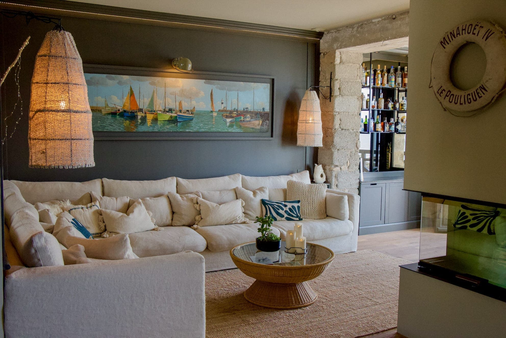 Un salon confortable avec un canapé d'angle beige et une table basse ronde en osier. La décoration nautique comprend un tableau de voilier et une bouée de sauvetage. De hautes lampes tissées ajoutent de la chaleur. Un bar bien approvisionné est visible en arrière-plan.