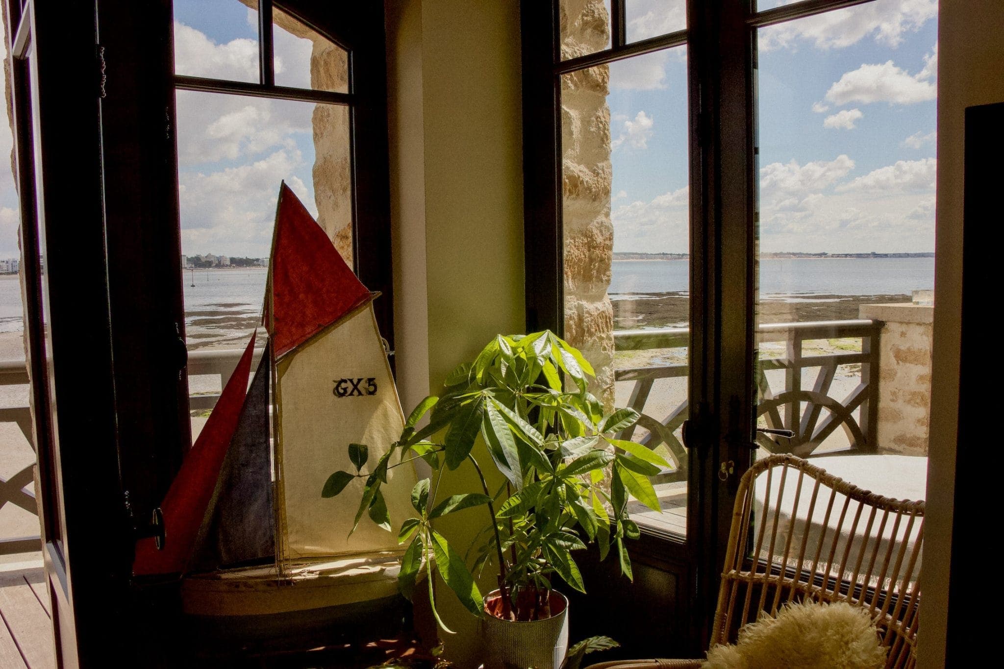 Une pièce confortable avec une grande fenêtre donnant sur une vue sur l'eau calme sous un ciel nuageux. À l'intérieur, il y a une maquette de voilier avec "GX3" imprimé dessus, une plante en pot et une chaise en osier. La lumière du soleil pénètre à flot, créant une atmosphère chaleureuse.