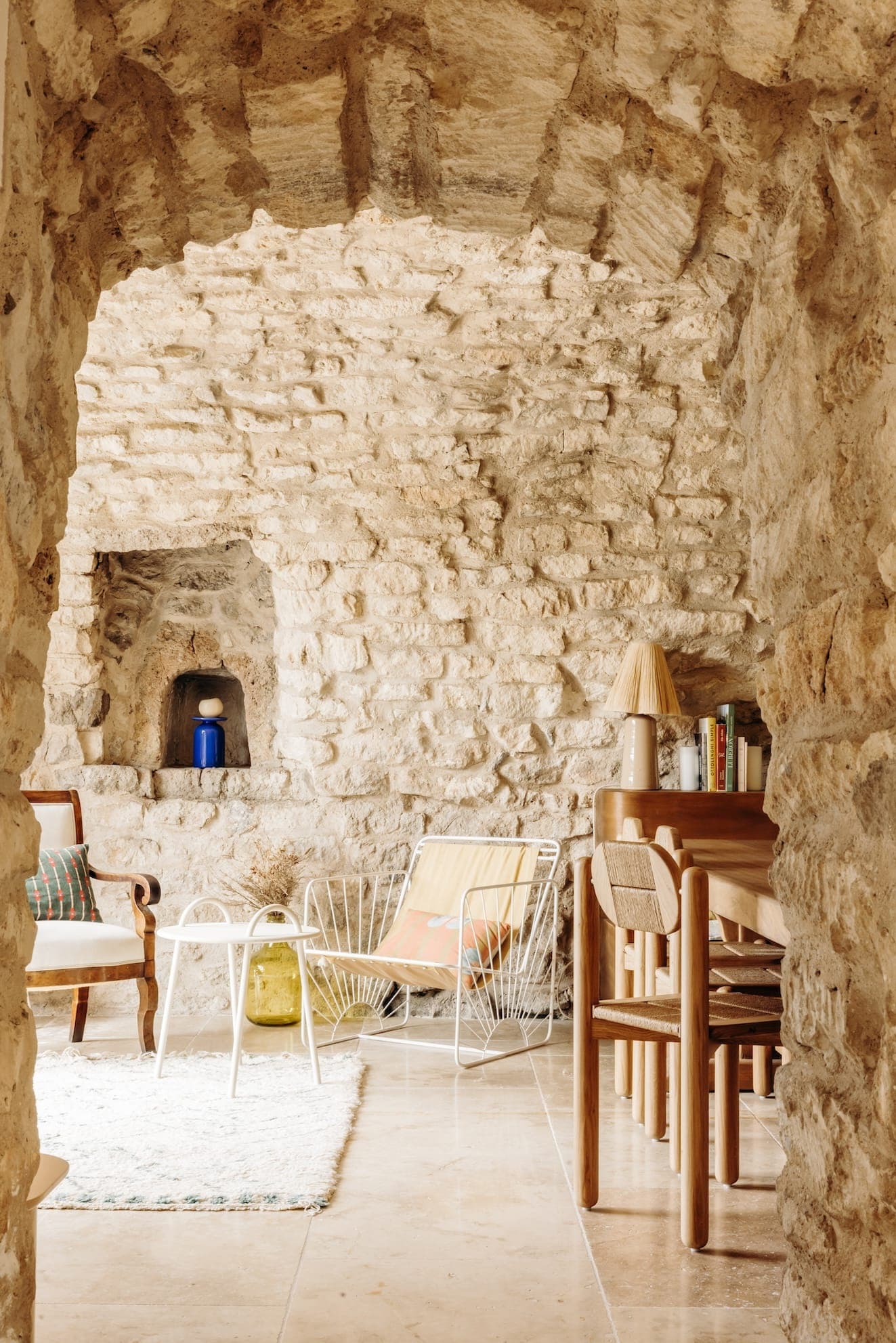 Une pièce rustique en pierre avec une entrée voûtée et des meubles modernes. On y trouve une table à manger en bois, des chaises, des coussins colorés et un tapis blanc. La lumière naturelle pénètre à flots, mettant en valeur une petite alcôve avec un vase bleu.