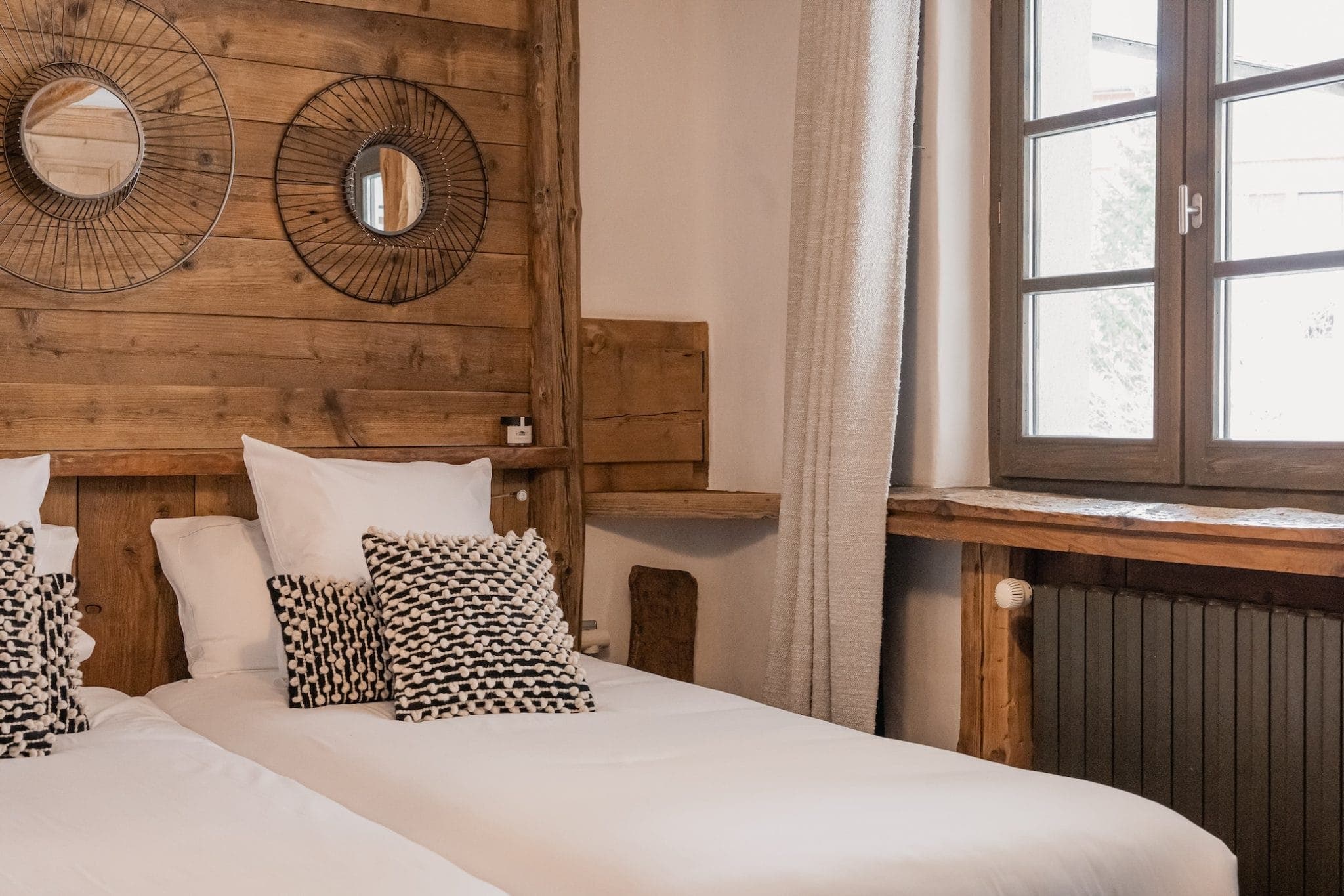 A cozy bedroom with two twin beds pushed together, featuring white bedding and patterned throw pillows. The walls are wooden, adorned with two round mirrors with intricate wire designs. A large window with beige curtains allows natural light to fill the room.