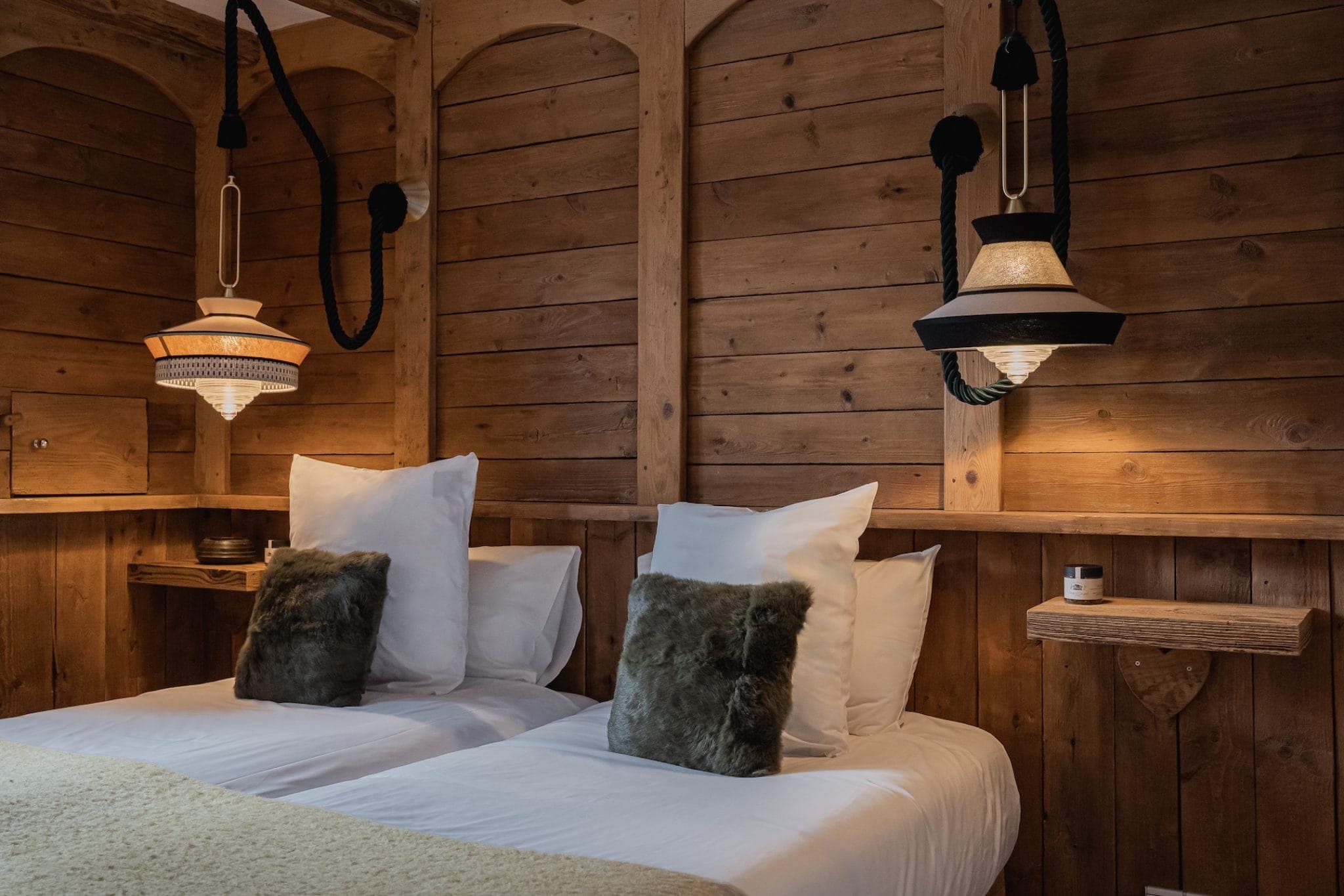 A cozy bedroom featuring two twin beds with white bedding and green throw pillows. The room has wooden paneled walls and rustic pendant lights hanging above each bed. A small built-in shelf on the wall holds a candle and other decorative items.