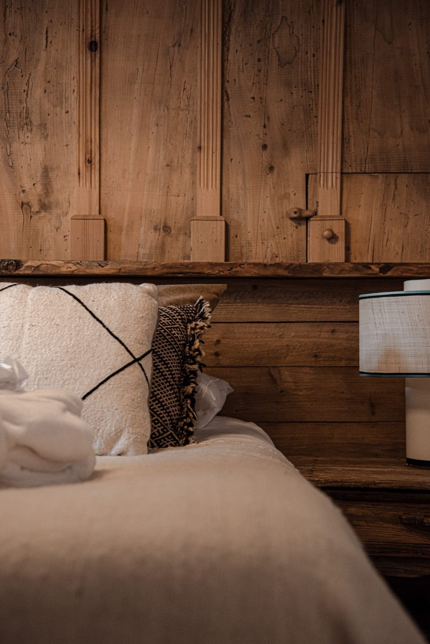 A cozy bedroom with a rustic wooden headboard and wall. The bed is adorned with white and patterned pillows, and there is a thick white blanket on it. A modern lamp with a beige shade sits on a wooden nightstand beside the bed.