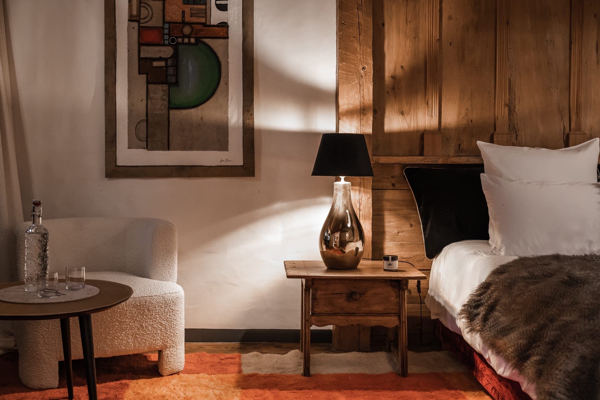 A cozy, warmly lit bedroom with rustic decor. A wooden bedside table holds a lamp with a black shade and a small cup. A white armchair and a round table with a bottle and glasses are on the left. A modern abstract painting hangs on the wall above the chair.