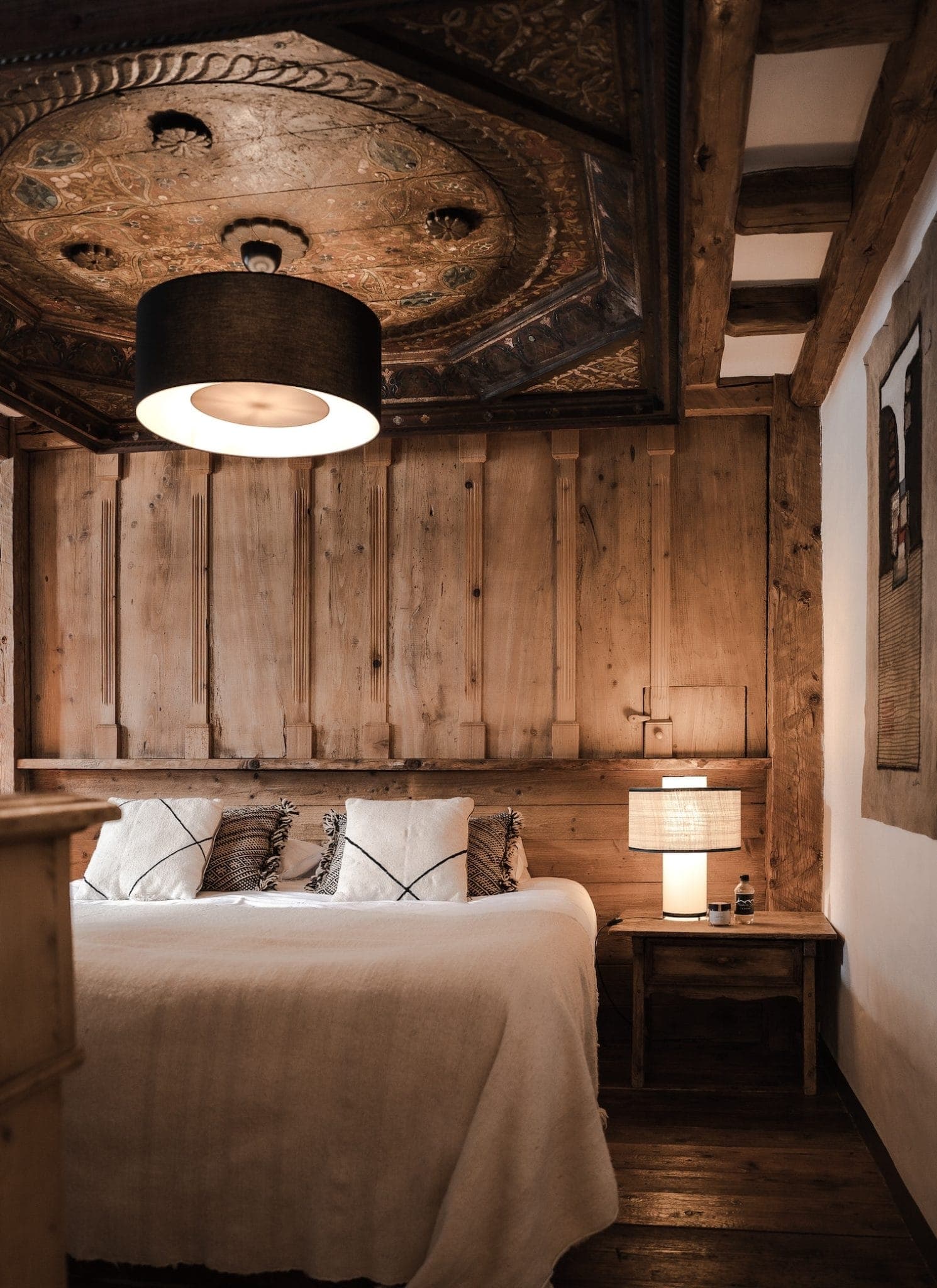 A rustic bedroom with wooden walls and ceiling features a large bed covered with white linens and several decorative pillows. A round, ornate ceiling medallion and a modern black pendant light hang above. A wooden nightstand with a lamp is beside the bed.