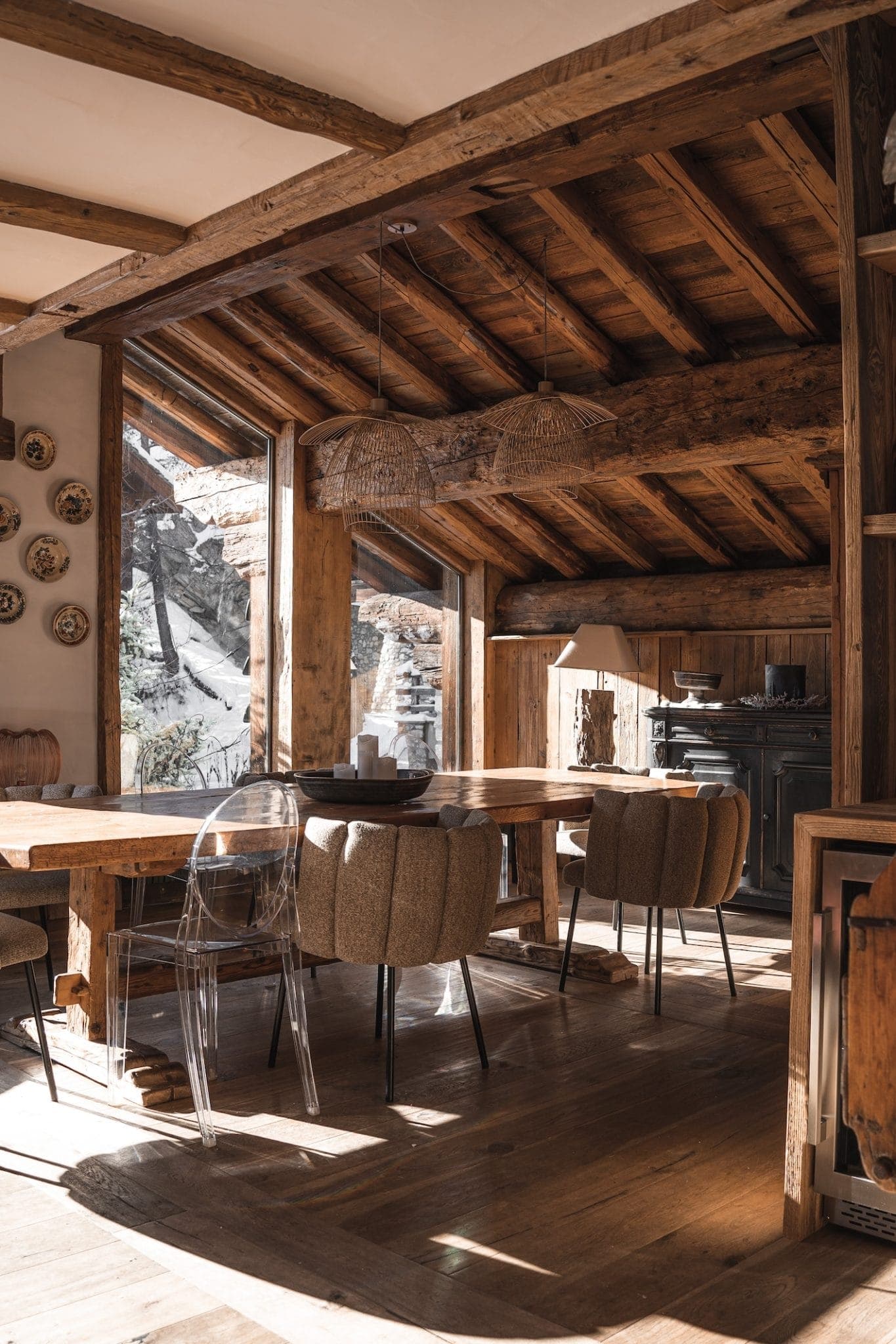 A rustic dining area with a wooden ceiling and exposed beams features a large wooden table surrounded by cushioned chairs and a transparent chair. Plates decorate one wall while large windows let in natural light, showcasing a snowy outdoor scene.