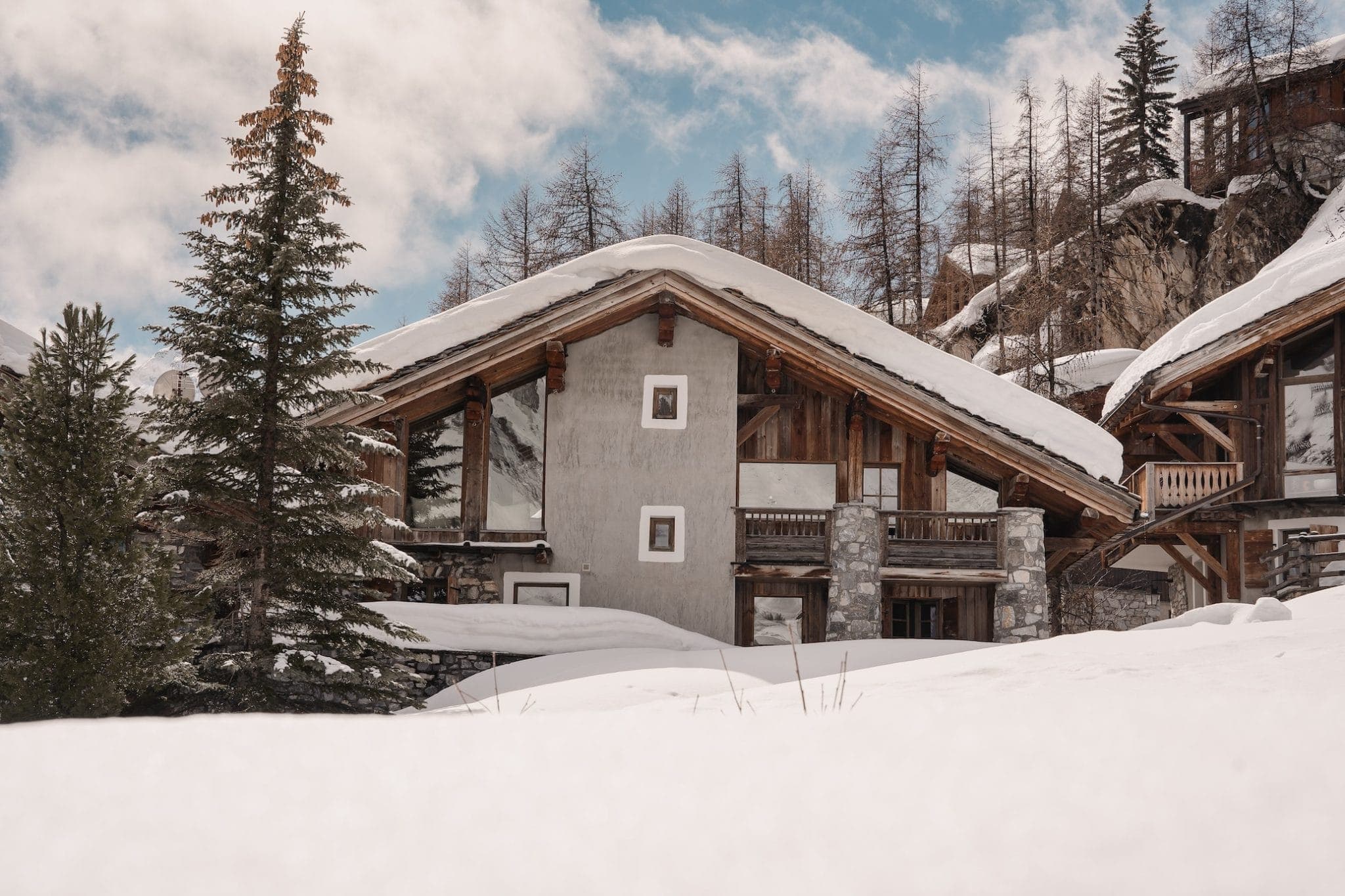 A cozy wooden cabin with a steep, snow-covered roof surrounded by trees and snow. The cabin has a rustic look with stone and wooden elements, large windows, and a balcony. The sky above is partly cloudy with patches of blue.