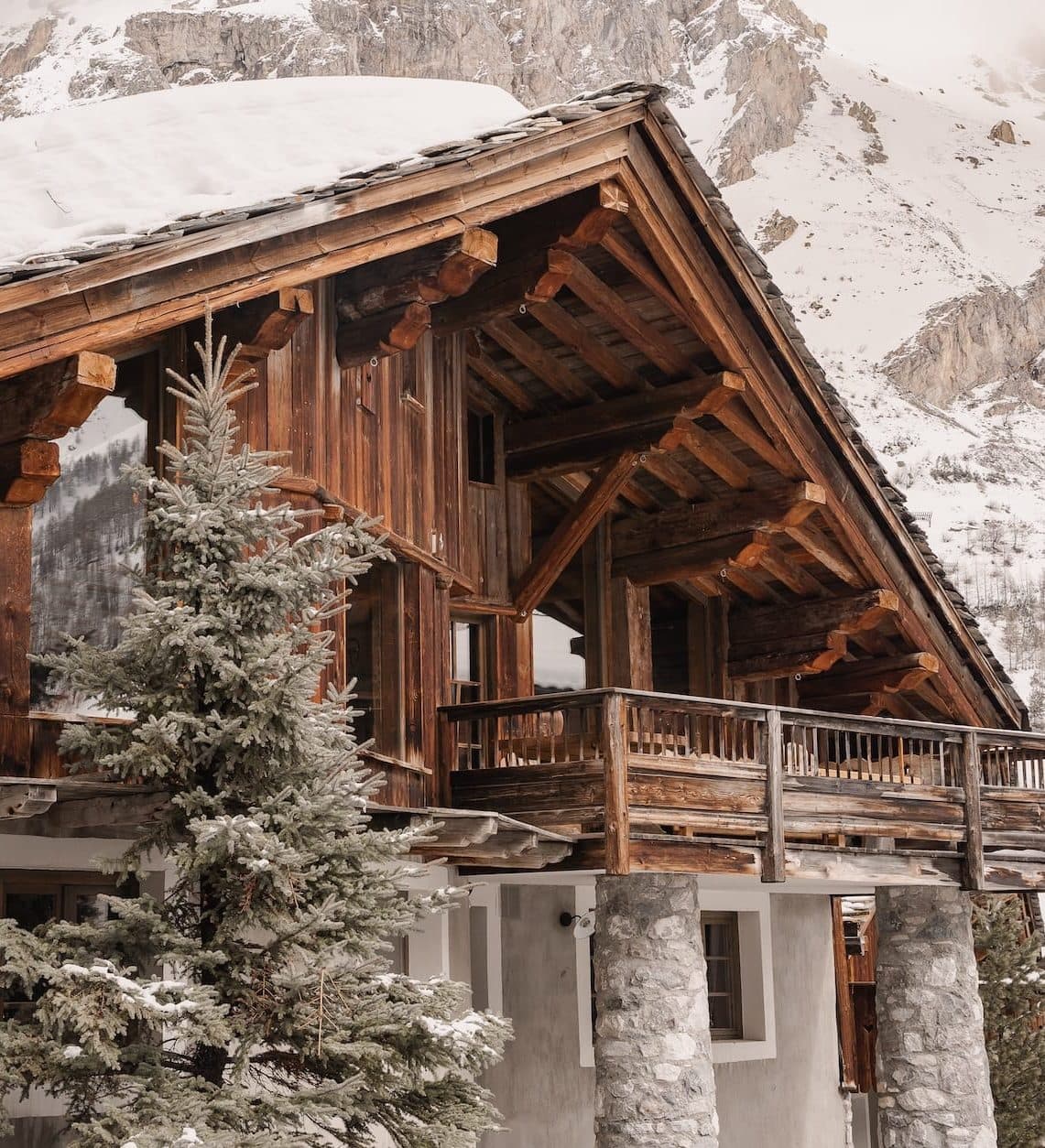 A rustic wooden cabin with stone pillars and a snow-covered roof is nestled among snow-laden pine trees against a backdrop of towering, snow-capped mountains. The scene evokes a cozy, serene winter setting.
