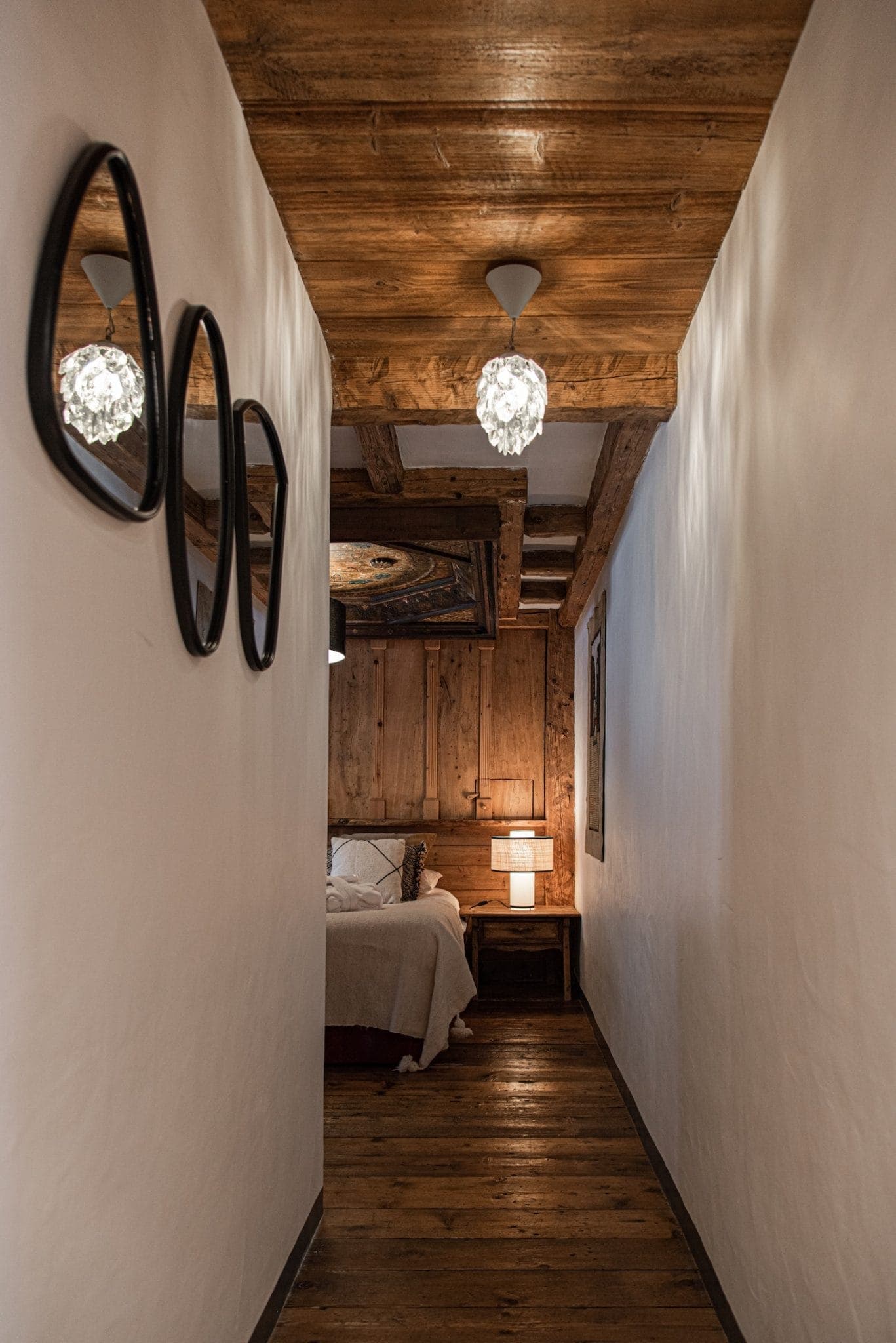 A narrow, rustic hallway with wooden ceiling beams leads to a cozy bedroom. The walls are adorned with a trio of oval mirrors on the left. The end of the hallway opens into a bedroom with a wooden headboard and a bedside lamp casting a warm glow.