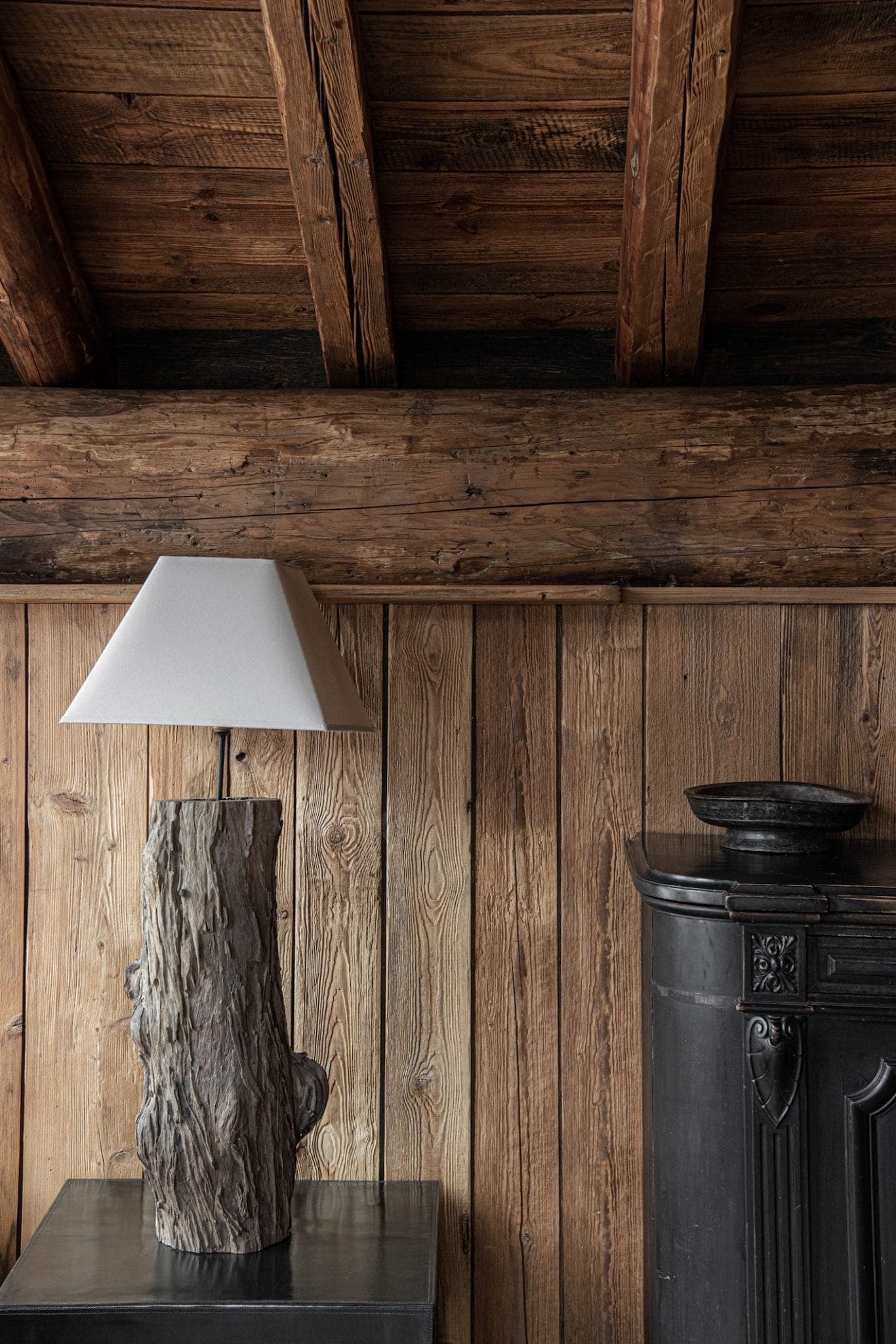 A rustic interior features a wooden table with a unique lamp made from a tree trunk and a white lampshade. The wood-paneled wall and exposed wooden beams complement the earthy tones. To the right, a dark, old-fashioned stove adds to the cozy ambiance.