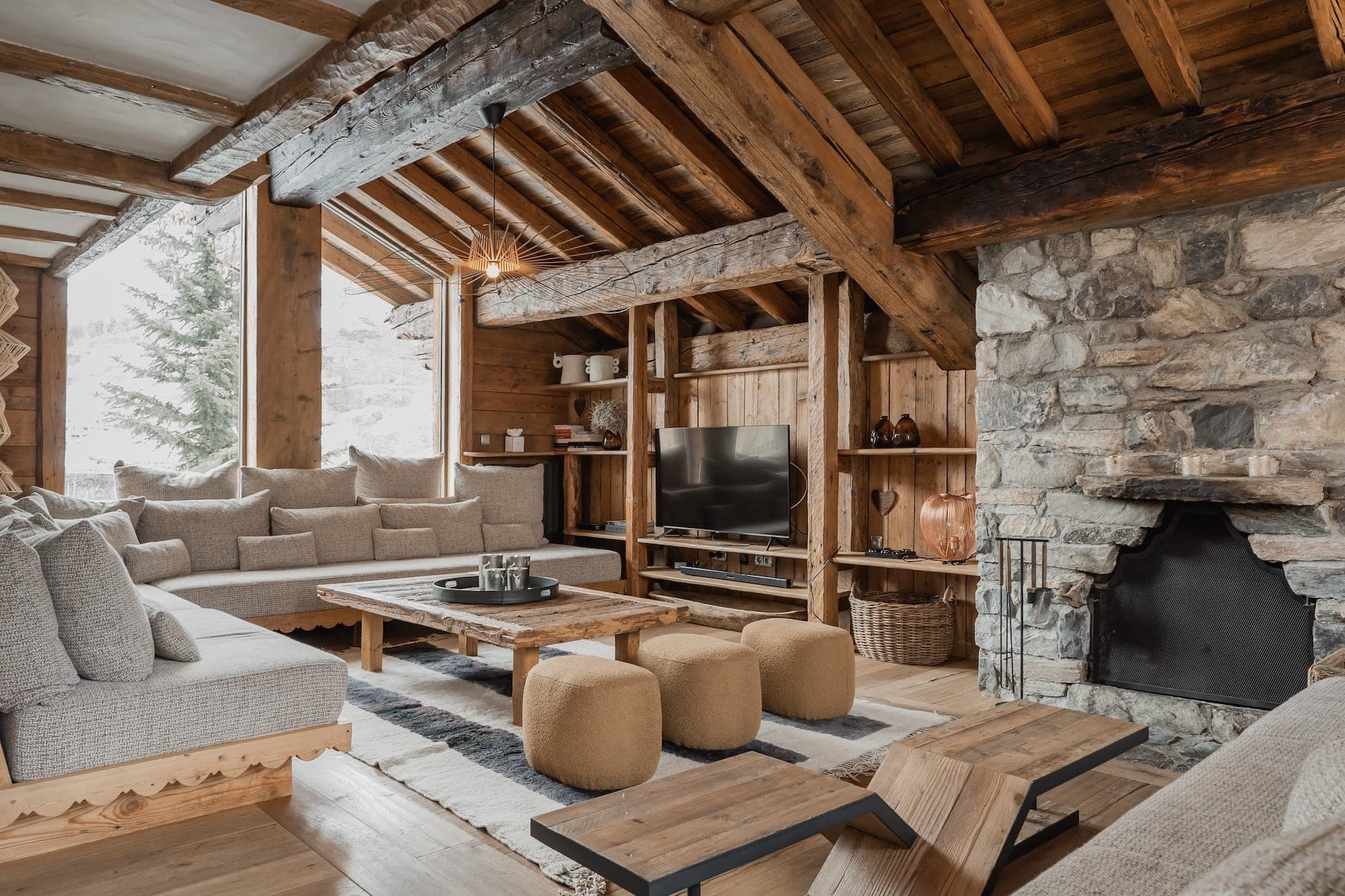 A cozy, rustic living room with wooden beams and ceilings, stone accents, a fireplace, and large windows. The room is furnished with beige sofas, a wooden coffee table, and two round poufs. There's a TV on a wooden shelf and a woven basket with firewood.