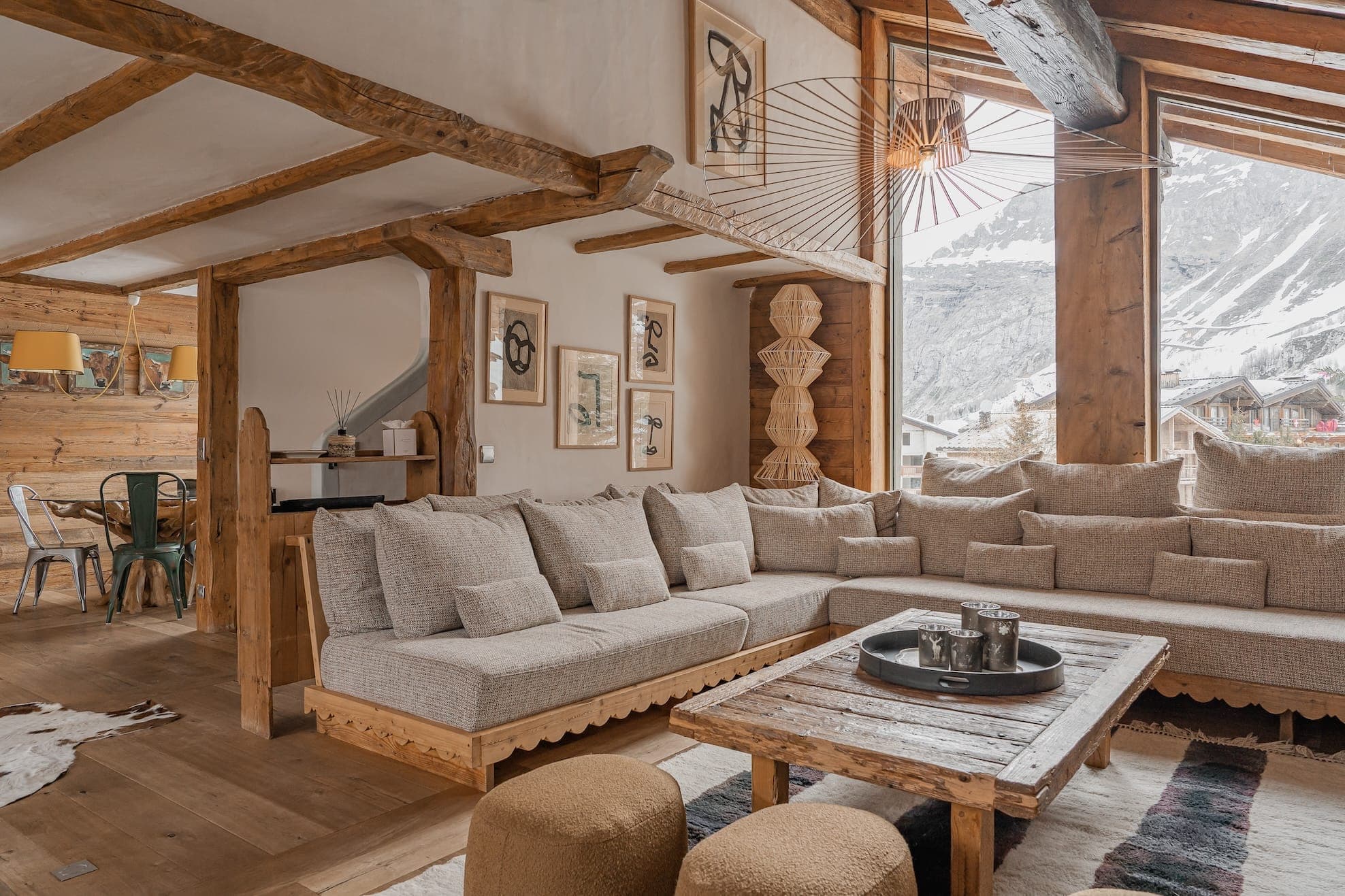 A cozy, rustic living room with large windows showcasing a snowy mountain view. The space features wooden beams, a beige sectional sofa, a wooden coffee table, and beige poufs. The room is decorated with modern art and wooden accents, creating a warm, inviting atmosphere.