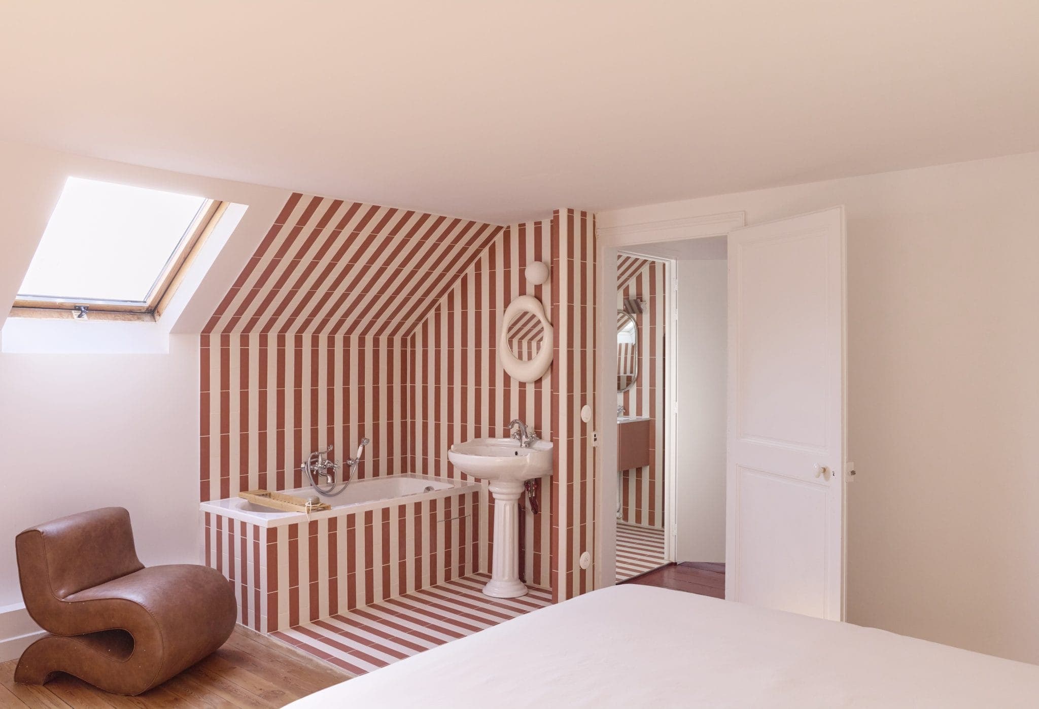 Une salle de bains moderne avec des carreaux géométriques à rayures rouges et blanches recouvrant les murs et le plafond en pente. Elle comprend une baignoire, un lavabo sur colonne, un miroir mural, une lucarne et une chaise marron unique. Une porte blanche partiellement ouverte révèle un couloir.