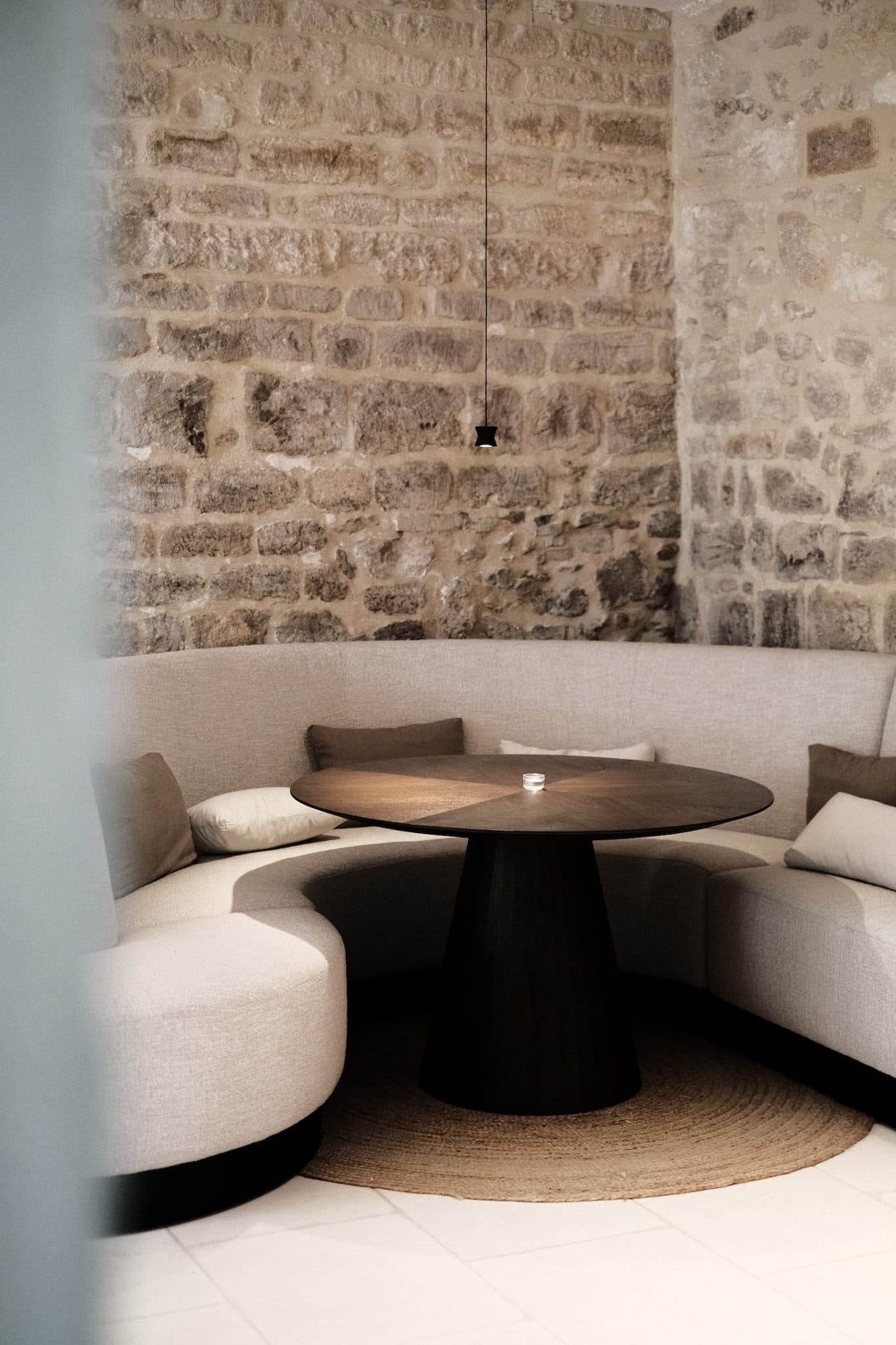 Un intérieur chaleureux et rustique comprend une table ronde en bois entourée d'un canapé beige incurvé, adossé à un mur en pierre texturé. Une petite suspension est suspendue au-dessus et des coussins ajoutent du confort au coin salon.