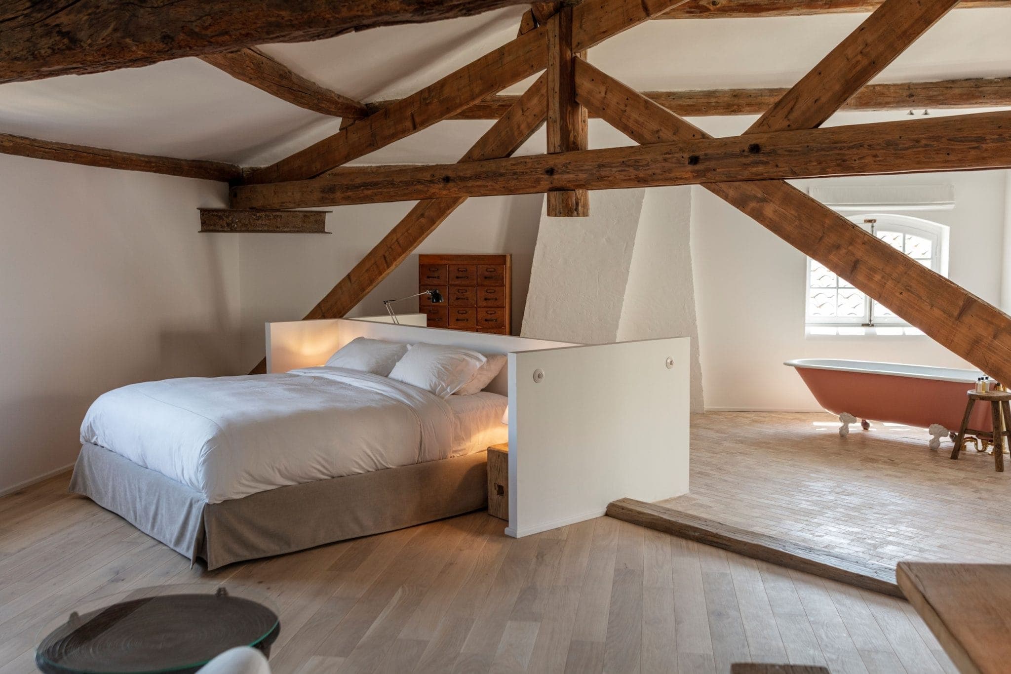 Une chambre mansardée confortable avec poutres apparentes en bois dispose d'un lit soigneusement fait avec des draps blancs. En arrière-plan, il y a une baignoire rose autoportante près d'une fenêtre. La chambre a du parquet et une décoration minimaliste.