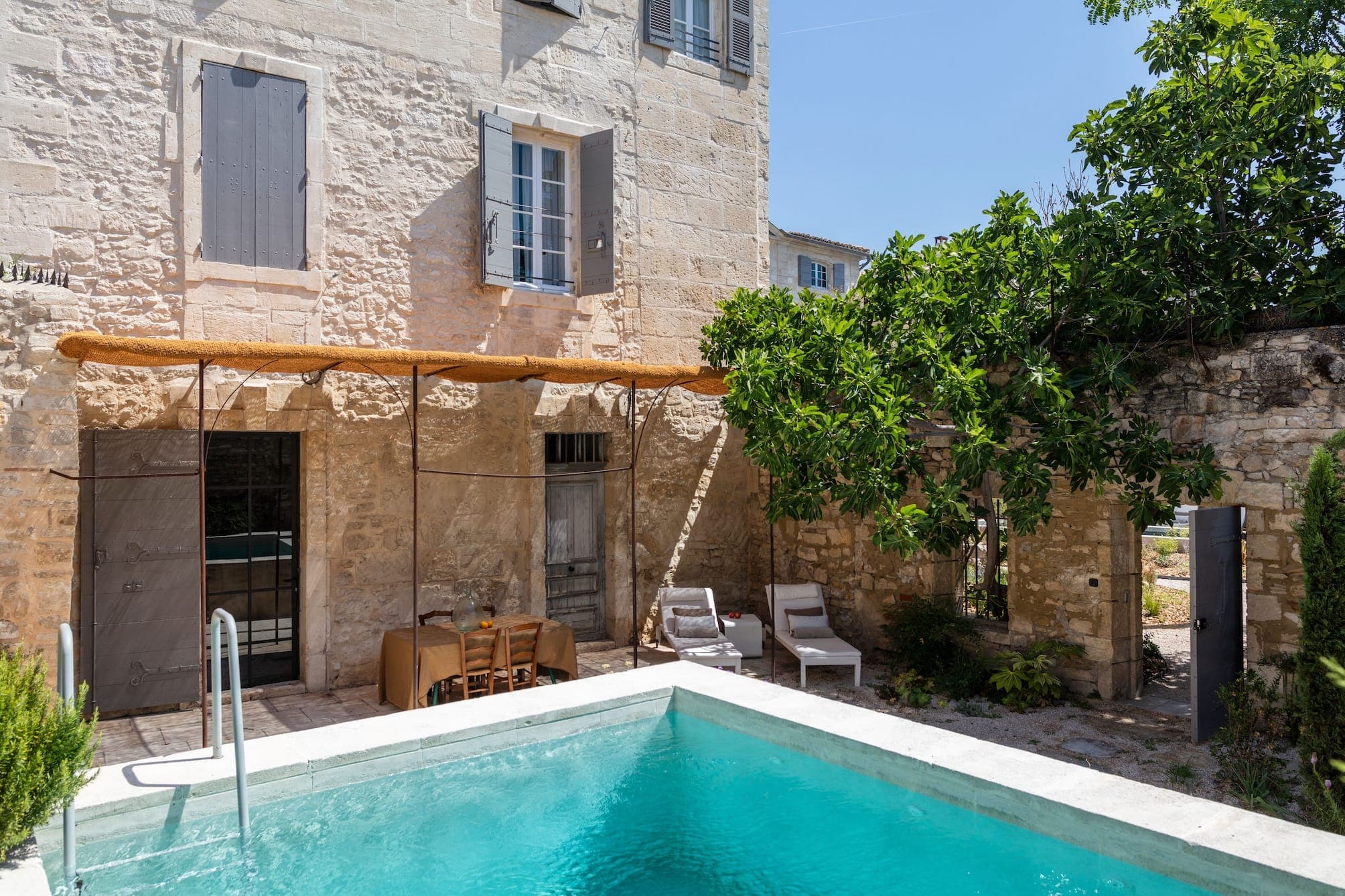 Une charmante maison en pierre aux volets en bois abrite une petite piscine au premier plan. Un patio ombragé avec une table à manger et des chaises à gauche et deux chaises longues à droite, entouré d'une végétation luxuriante et d'un grand arbre.