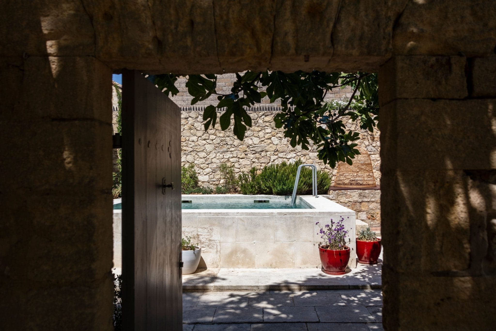 Une vue à travers une porte en pierre révèle une piscine extérieure tranquille entourée de murs en pierre. Des plantes en pot, notamment des fleurs violettes et rouges, décorent l'espace, et des branches feuillues pendent au-dessus. La scène est baignée de lumière naturelle.