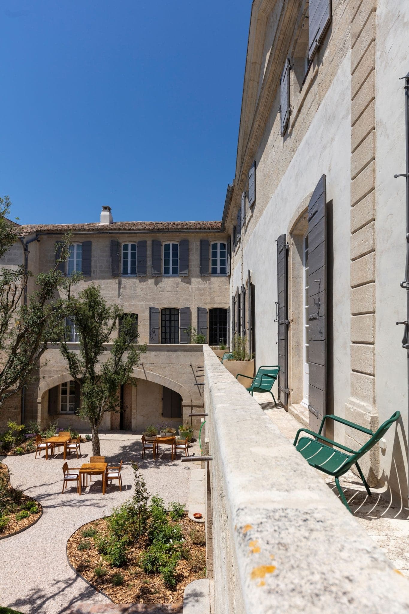 Une cour d'un bâtiment historique avec un jardin rond avec de petits arbres, entouré de gravier et de chaises pour s'asseoir. La façade comprend des volets sur les fenêtres et les portes. Un ciel bleu clair est visible au-dessus.