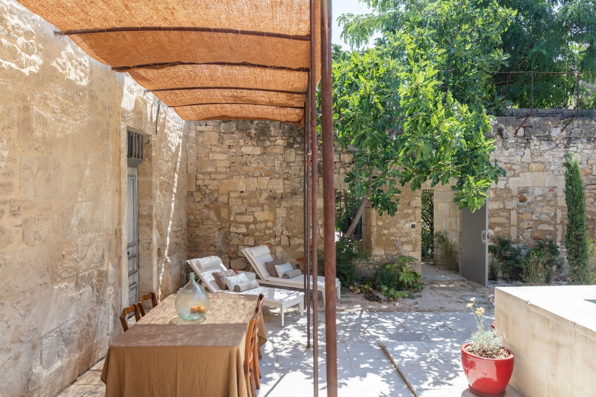 Une cour ensoleillée avec un mur en pierre et un feuillage vert, comprenant un coin repas extérieur ombragé avec une table et des chaises recouvertes de tissu. À proximité se trouvent deux chaises longues et une plante en pot dans un pot rouge. Une porte en bois est encastrée dans le mur.