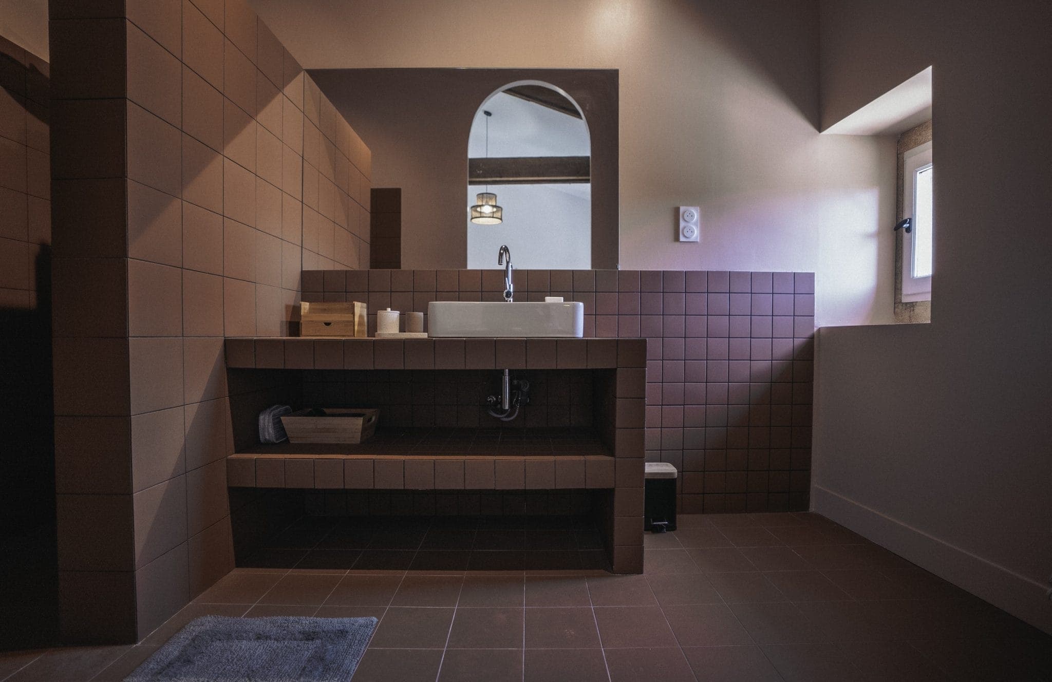 A bathroom with brown tiled walls and floor. The space features a white sink with a large mirror above it, shelves underneath the sink holding various toiletries, and a small window letting in natural light. A gray rug lies on the floor in front of the sink.
