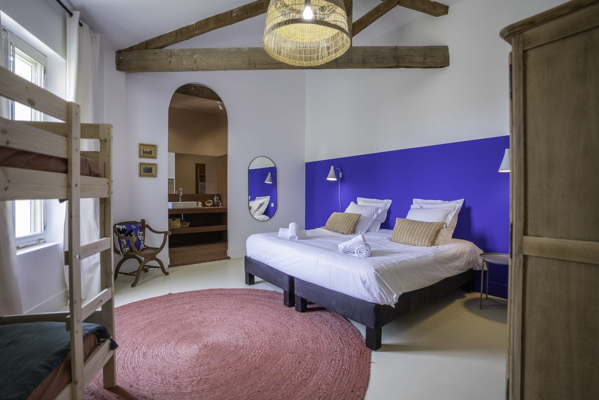 A cozy bedroom with a double bed and bunk bed. The room features a vibrant blue accent wall, a red circular rug, wooden ceiling beams, and a wicker lampshade. A wooden wardrobe, an arched mirror, and a small armchair complete the space.