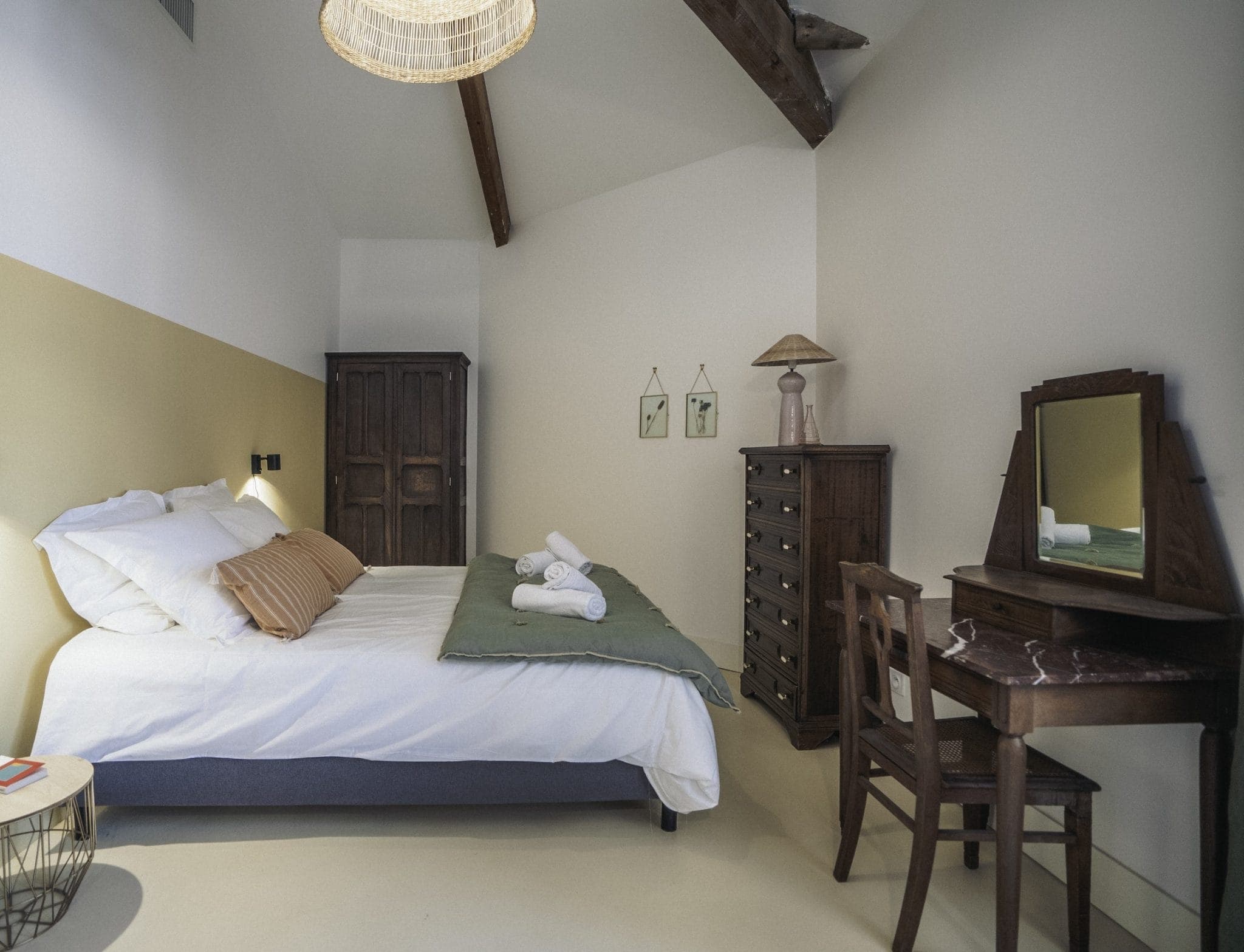 A cozy bedroom features a bed with white linens and peach pillows, neatly folded towels, a wooden wardrobe, and a matching wooden dresser. A wooden vanity with a mirror and chair stands by the wall. Warm lighting and exposed ceiling beams complete the rustic look.